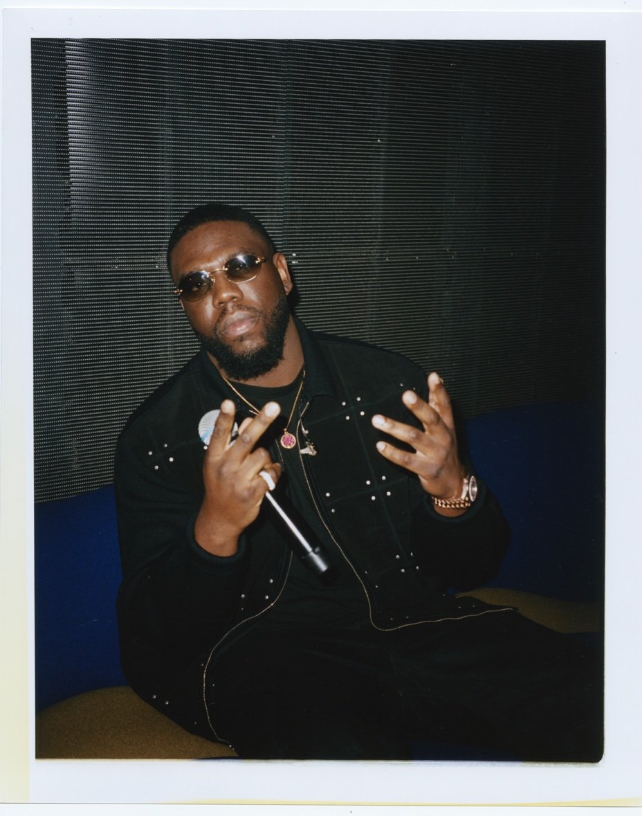 Un homme portant des lunettes de soleil, une veste noire avec des détails brillants, tenant un microphone et faisant un signe de paix avec ses doigts. Assis sur un canapé bleu devant un mur en panneaux noirs.