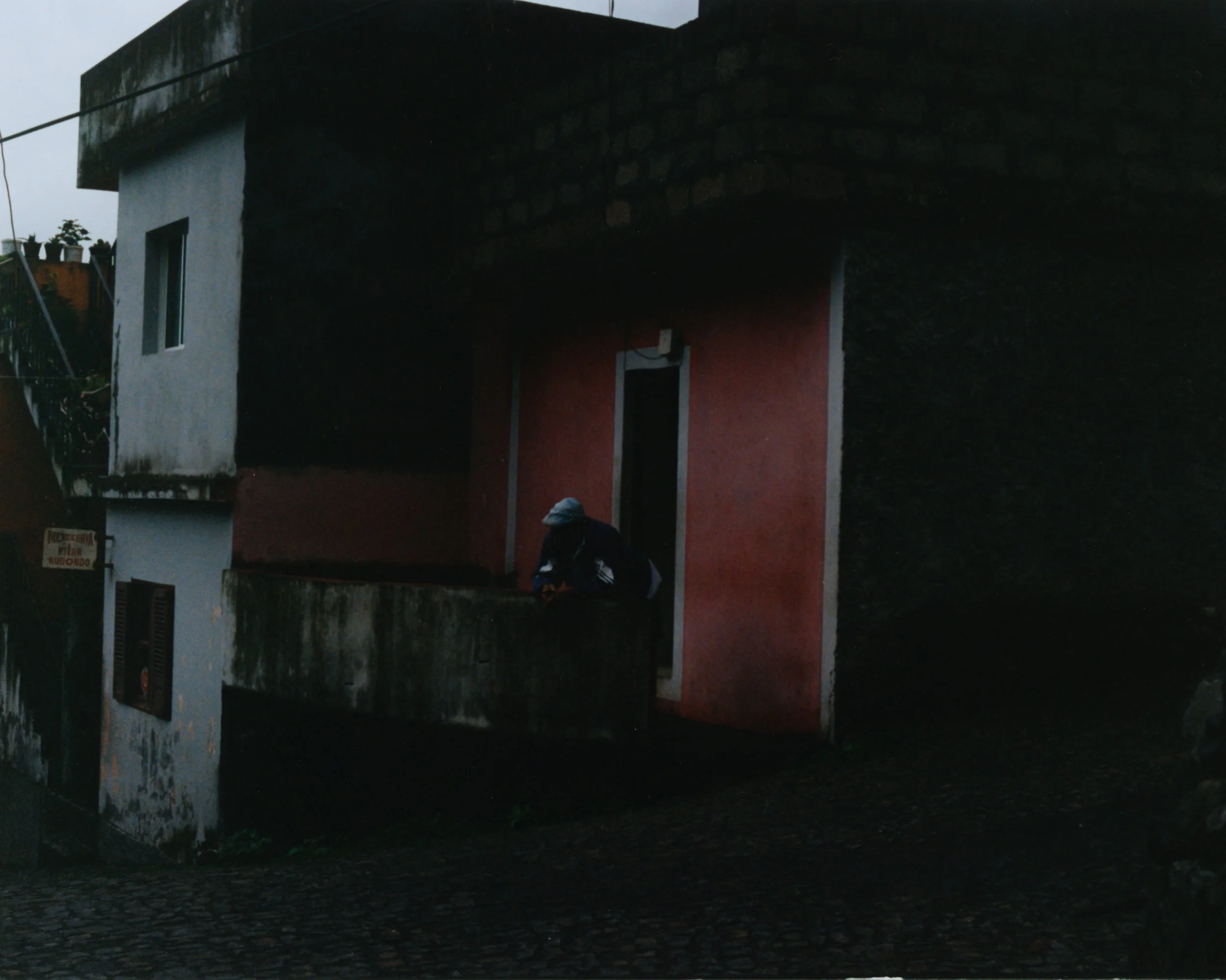 Une maison avec deux étages, une partie blanche et une partie rouge, un homme porte une casquette à genoux près d'une balustrade en béton, dans une zone résidentielle.