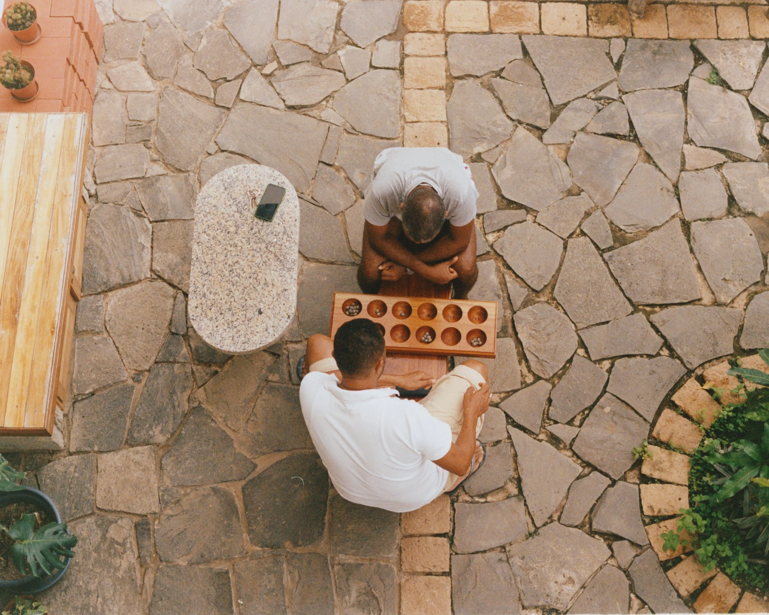 Deux personnes jouent à un jeu de société sur une terrasse pavée. L'une est assise, l'autre est accroupie, et un plateau de jeu est posé entre eux.