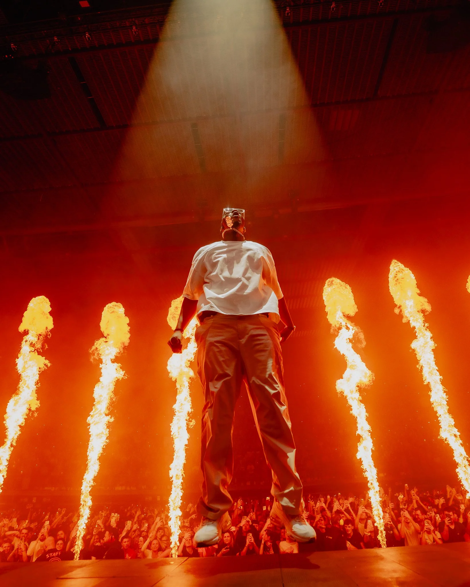Un artiste en scène lors d'un concert avec des flammes en arrière-plan, portant une chemise blanche et des pantalons clairs, le public en dessous.
