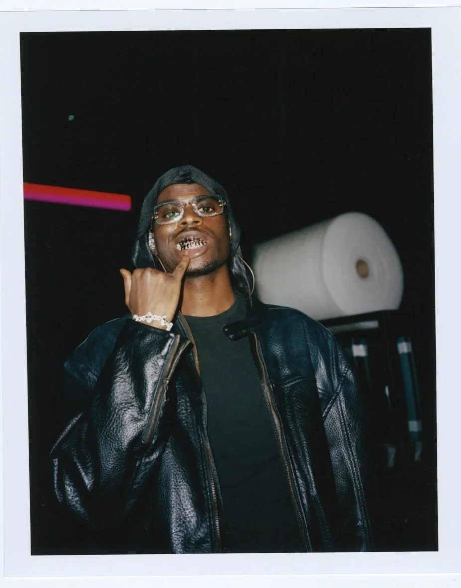 Homme portant des lunettes et un bustier en cuir noir avec des tatouages en or sur les dents, faisant un geste avec la main dans un environnement sombre avec un rouleau de papier en arrière-plan.