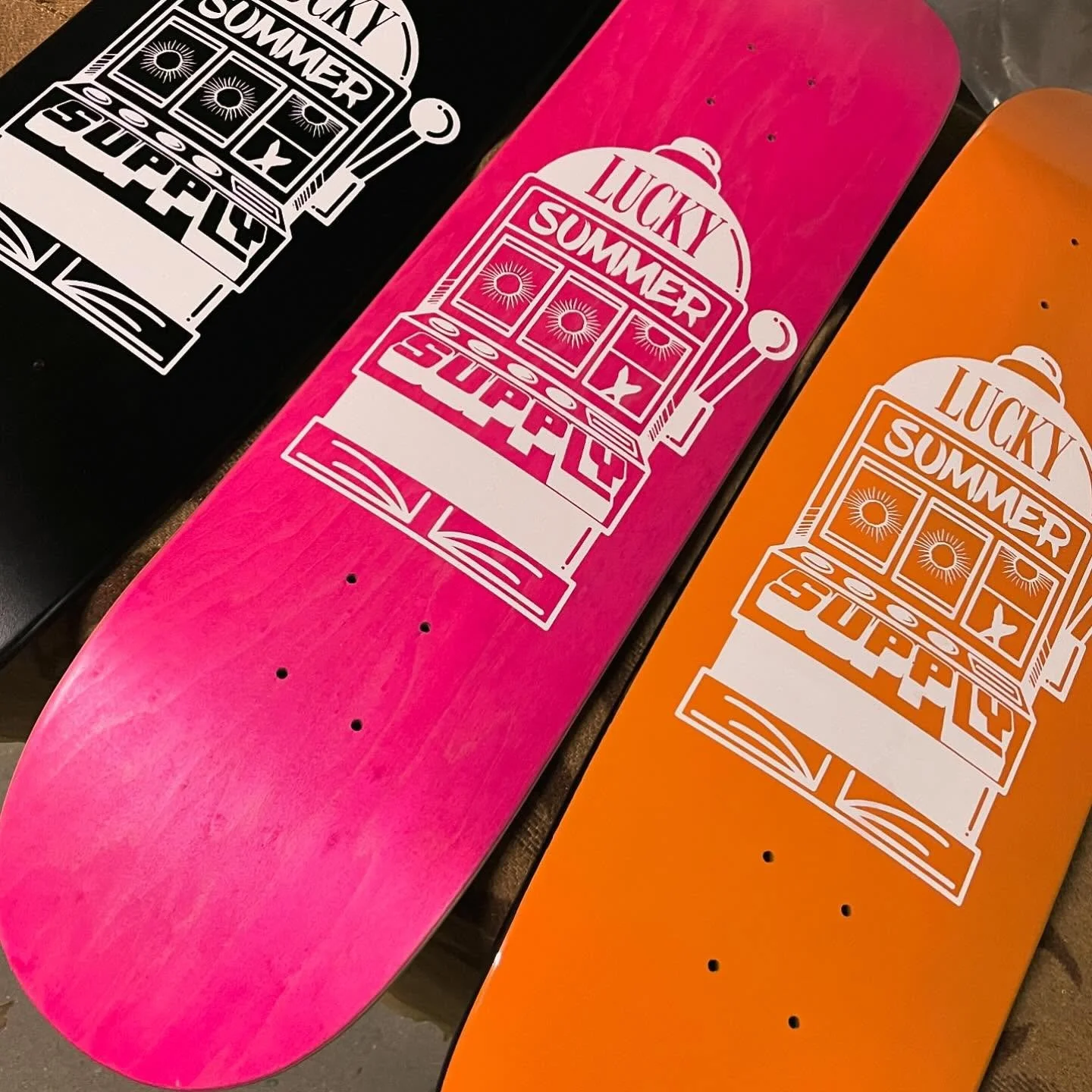 Drei Skateboards mit bunten Decks, eines in Schwarz, eines in Pink und eines in Orange, alle mit einem weißen Aufdruck eines Slot-Maschine und dem Text "Lucky Summer" und "Super Luck".