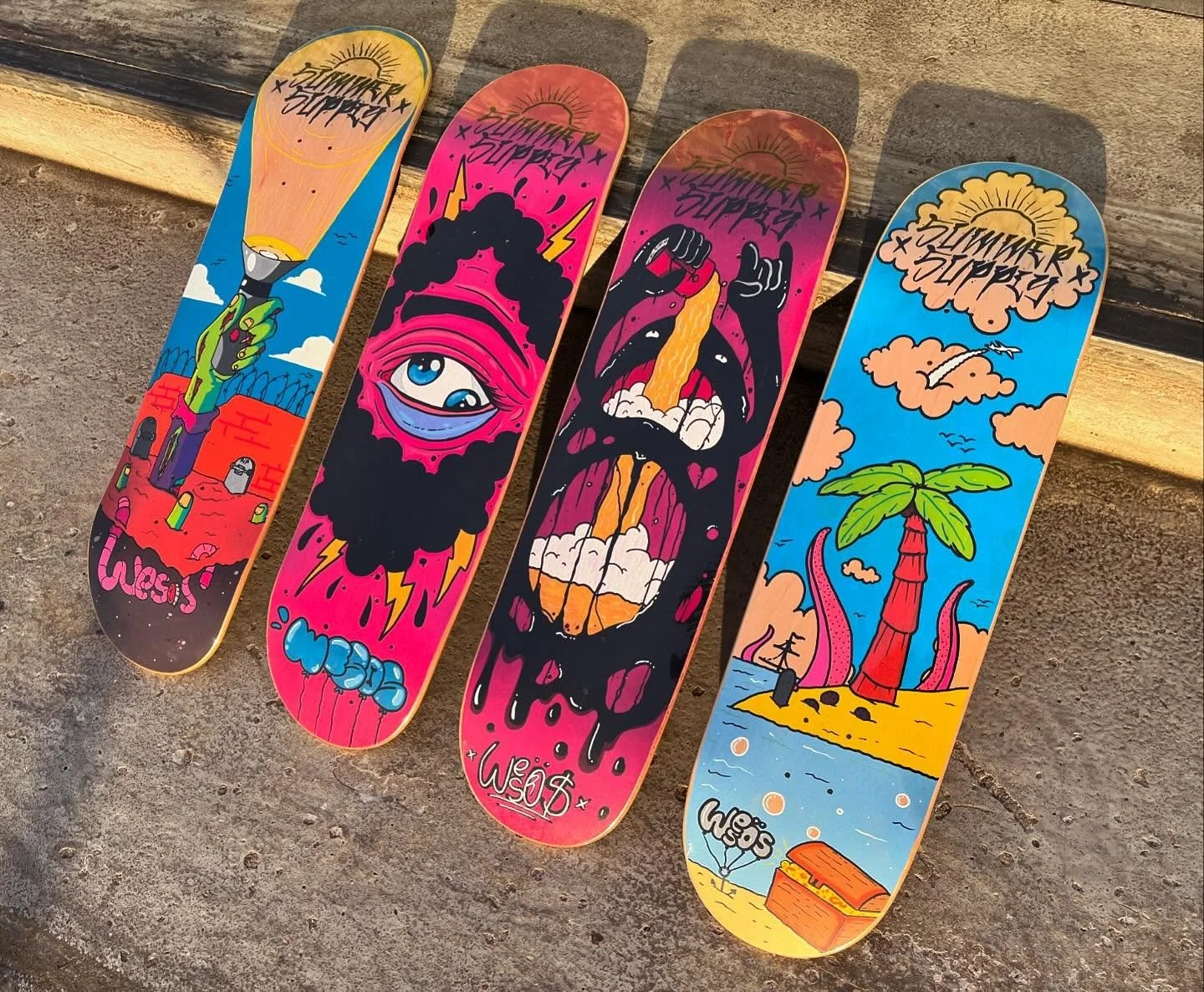 Vier Skateboards mit bunten, auffälligen Grafiken im Skatepark. Das erste ist eine Hand, die einen Rauchmelder hält, das zweite zeigt ein Auge mit Wolken und Blitzen, das dritte eine aufgerissene Mund mit einer Zunge und den Text 'Lust' und 'Wos', und das vierte zeigt eine Insel mit Palmen und einem Schatz im Sand.