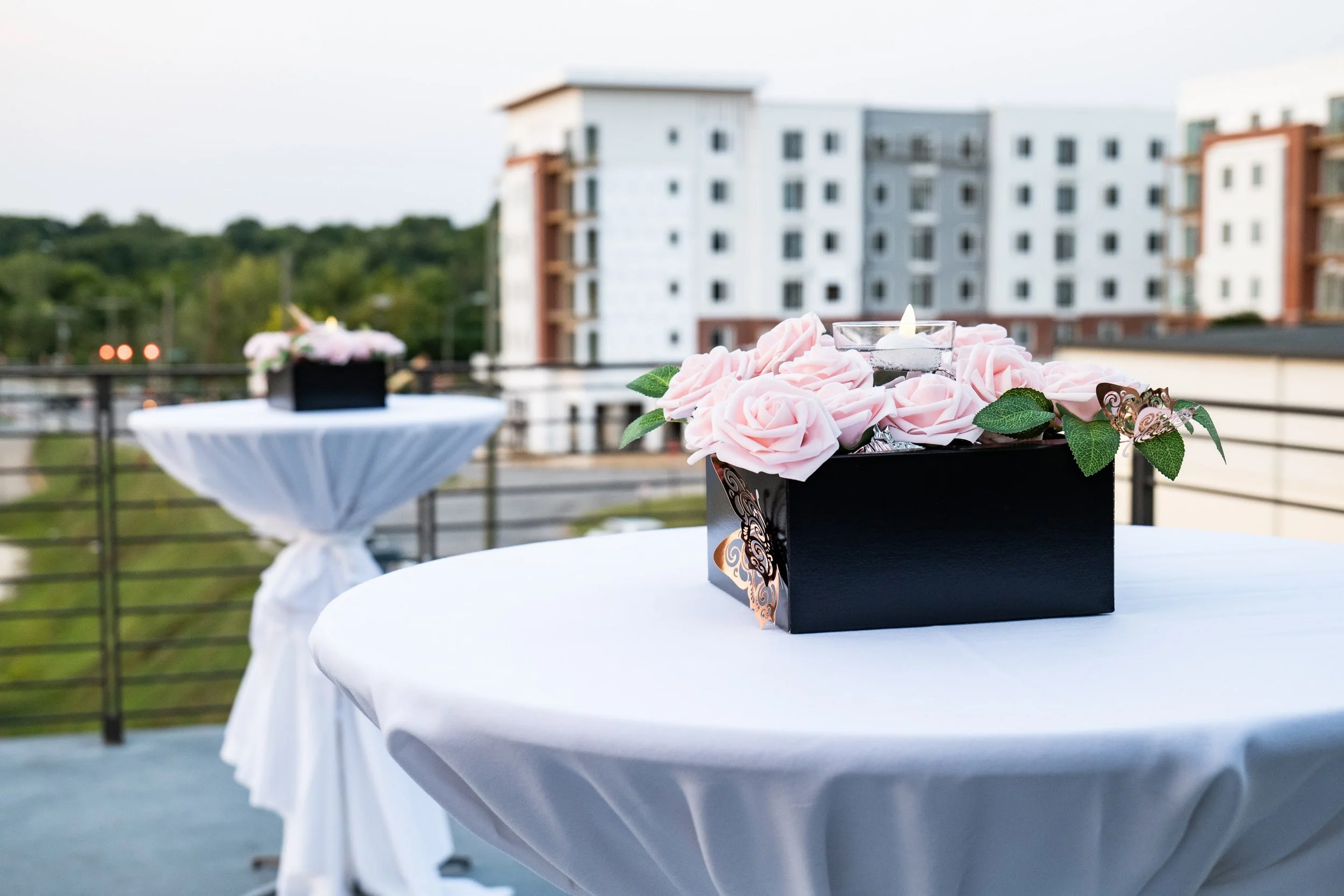 A black gift box with pink artificial roses inside, sitting on a white tablecloth-covered table on an outdoor balcony with a view of a modern apartment building and green trees in the background.
