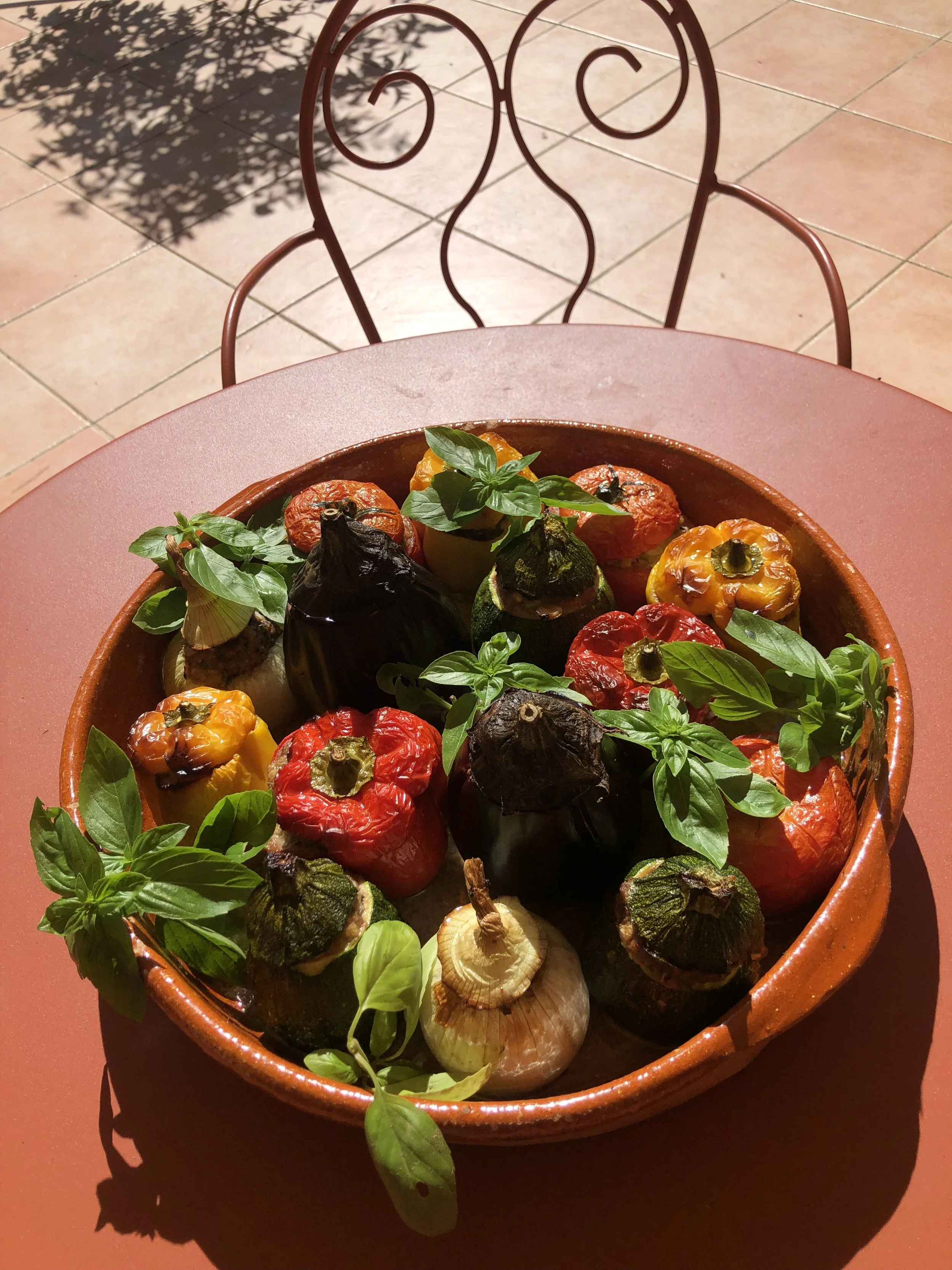 Un plat de poivrons, aubergines, tomates, échalotes et basilic frais dans un plat en céramique, avec une table en fer forgé en arrière-plan.