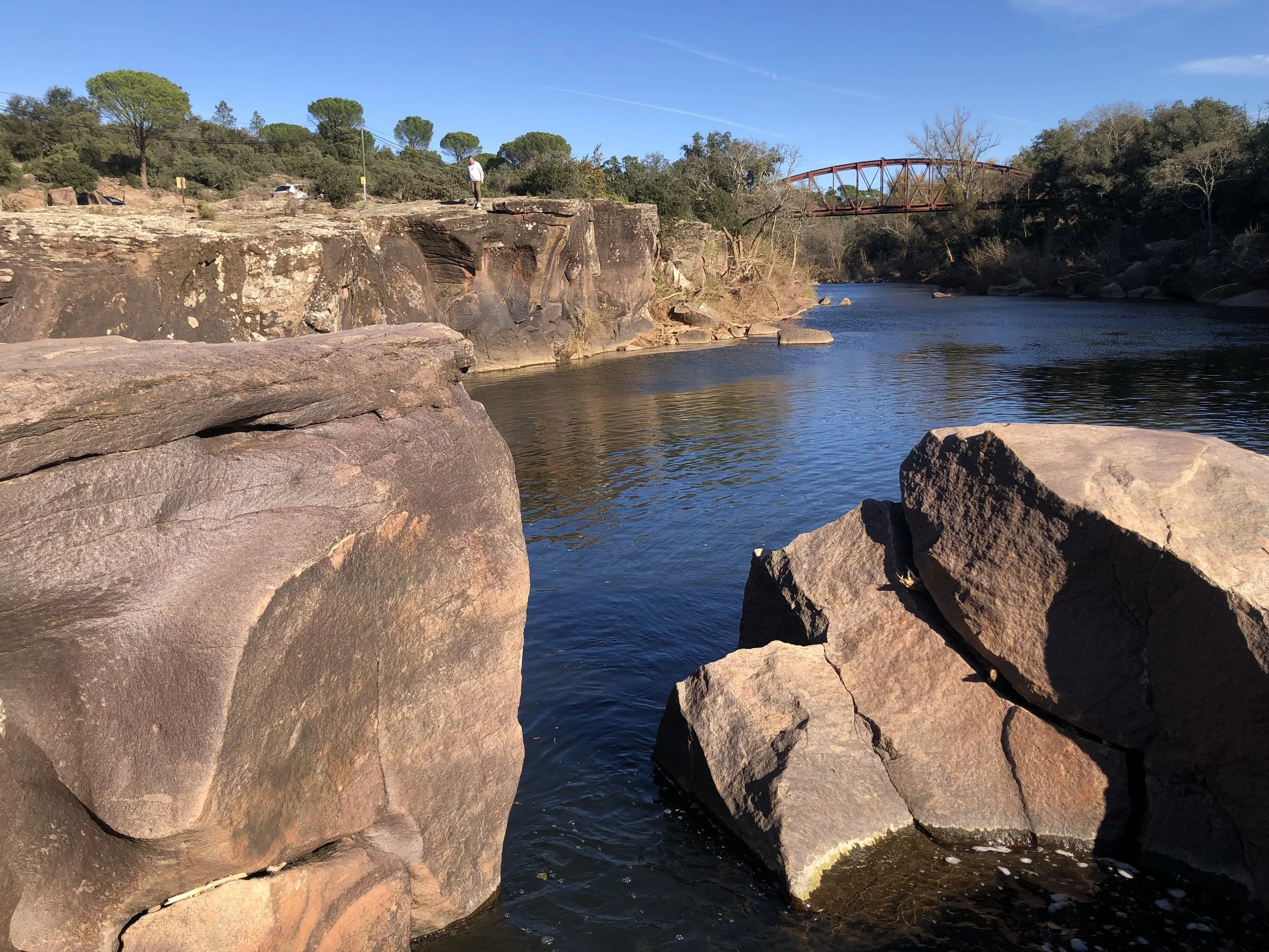 Rivière bordée de rochers sous un ciel bleu avec des arbres et un pont métallique en arrière-plan.