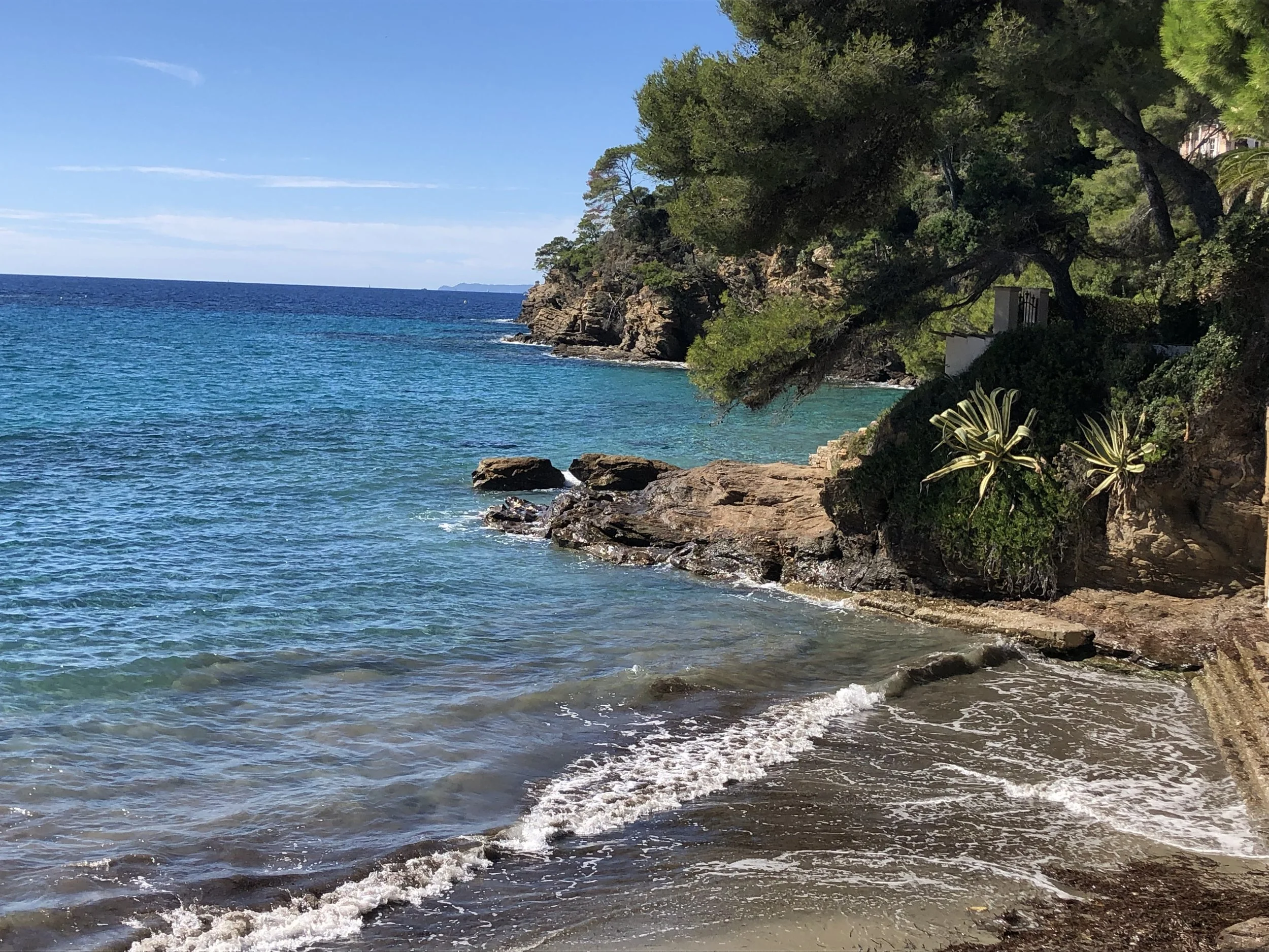 Plage rocheuse avec vaguelette, eau turquoise, arbres verts et rocheux, ciel bleu.