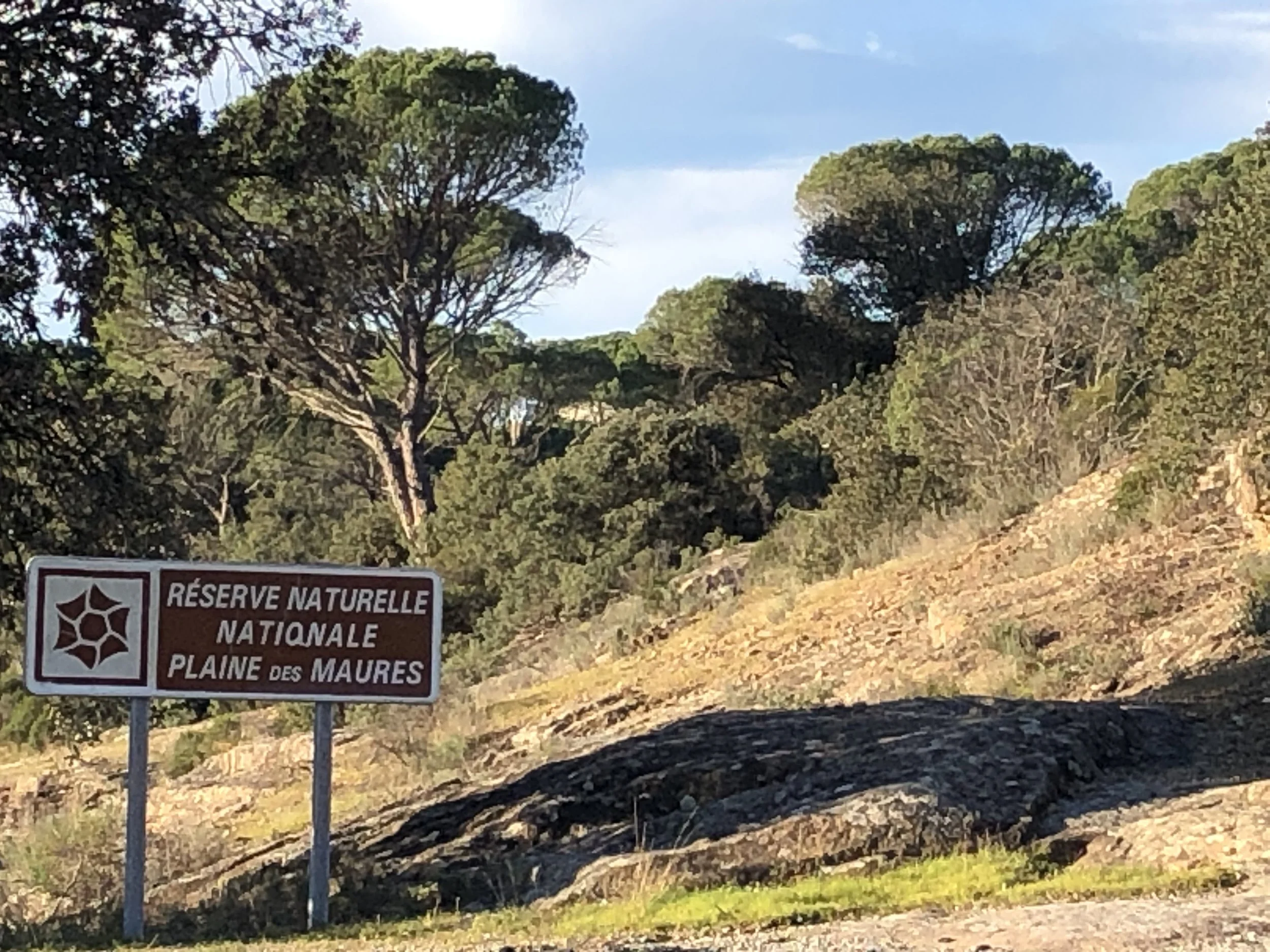 Un panneau marron indiquant 'Réserve Naturelle Nationale Plaine des Maures' avec un symbole d'araignée, dans un paysage rocheux avec des arbres verts et un ciel bleu.