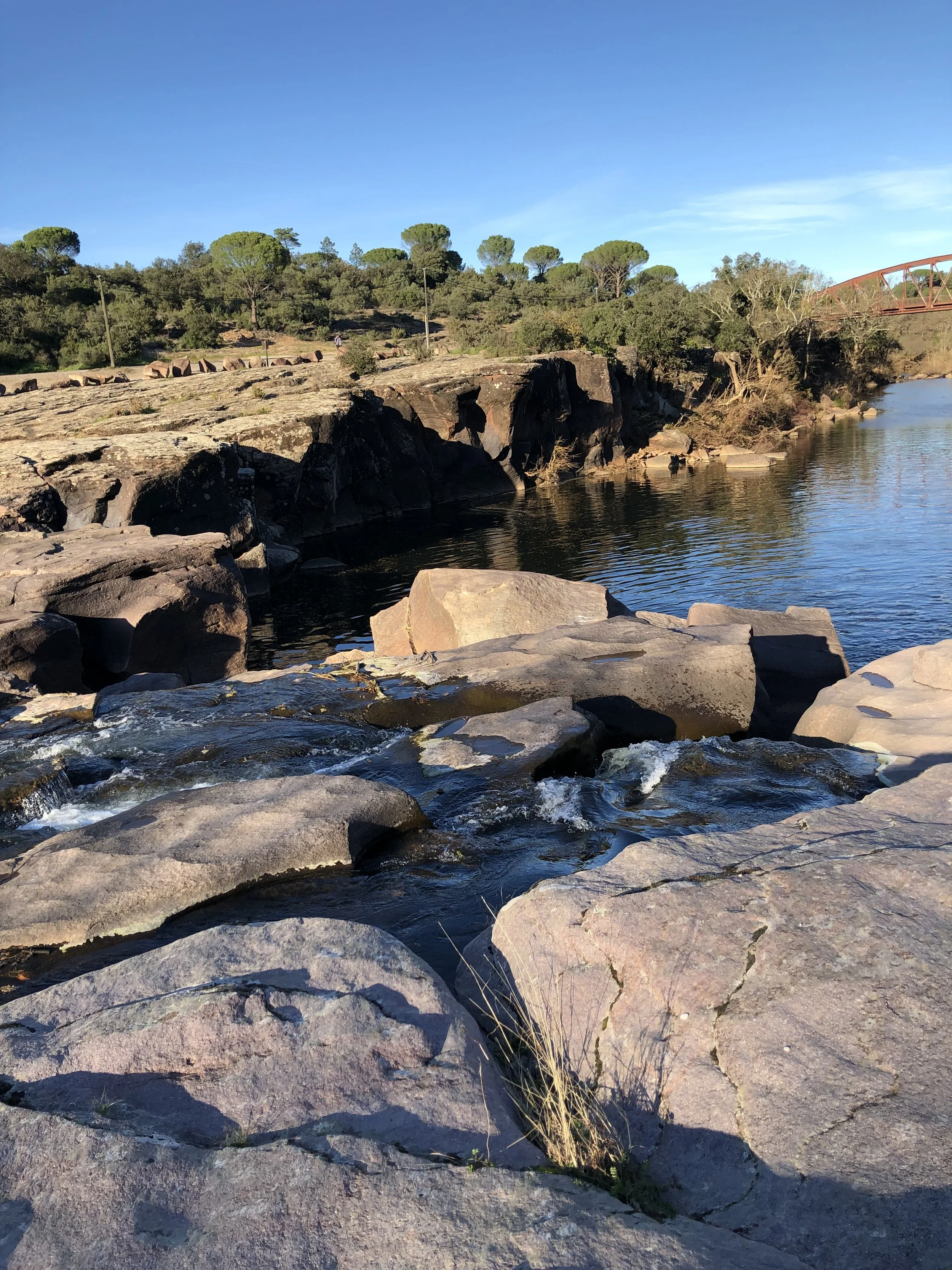 Roches au bord d'une rivière avec des arbres et un pont en métal en arrière-plan sous un ciel bleu.
