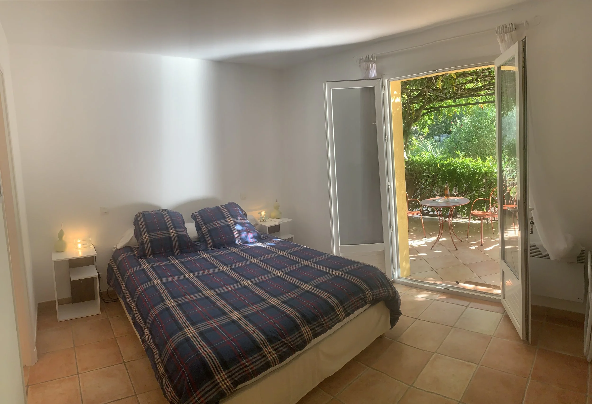 Chambre avec un lit double au drap à carreaux, deux tables de nuit avec lampes, porte-fenêtre donnant sur un balcon avec petit salon extérieur, vue sur un jardin verdoyant