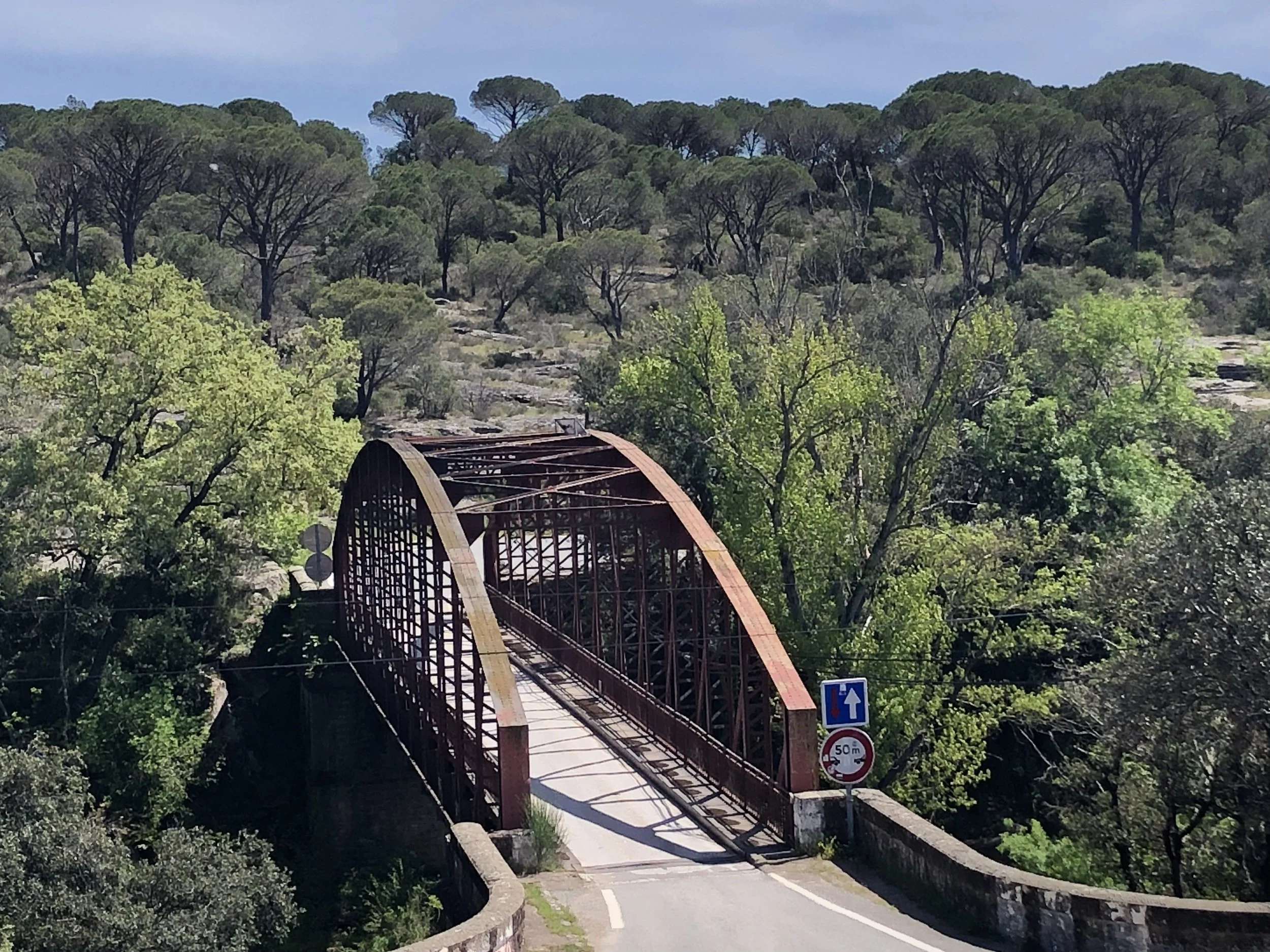 Un vieux pont en métal rouge qui enjambe une petite route, entouré d'arbres verts et de collines boisées, avec un panneau de signalisation indiquant une hauteur maximale de 50 mètres.