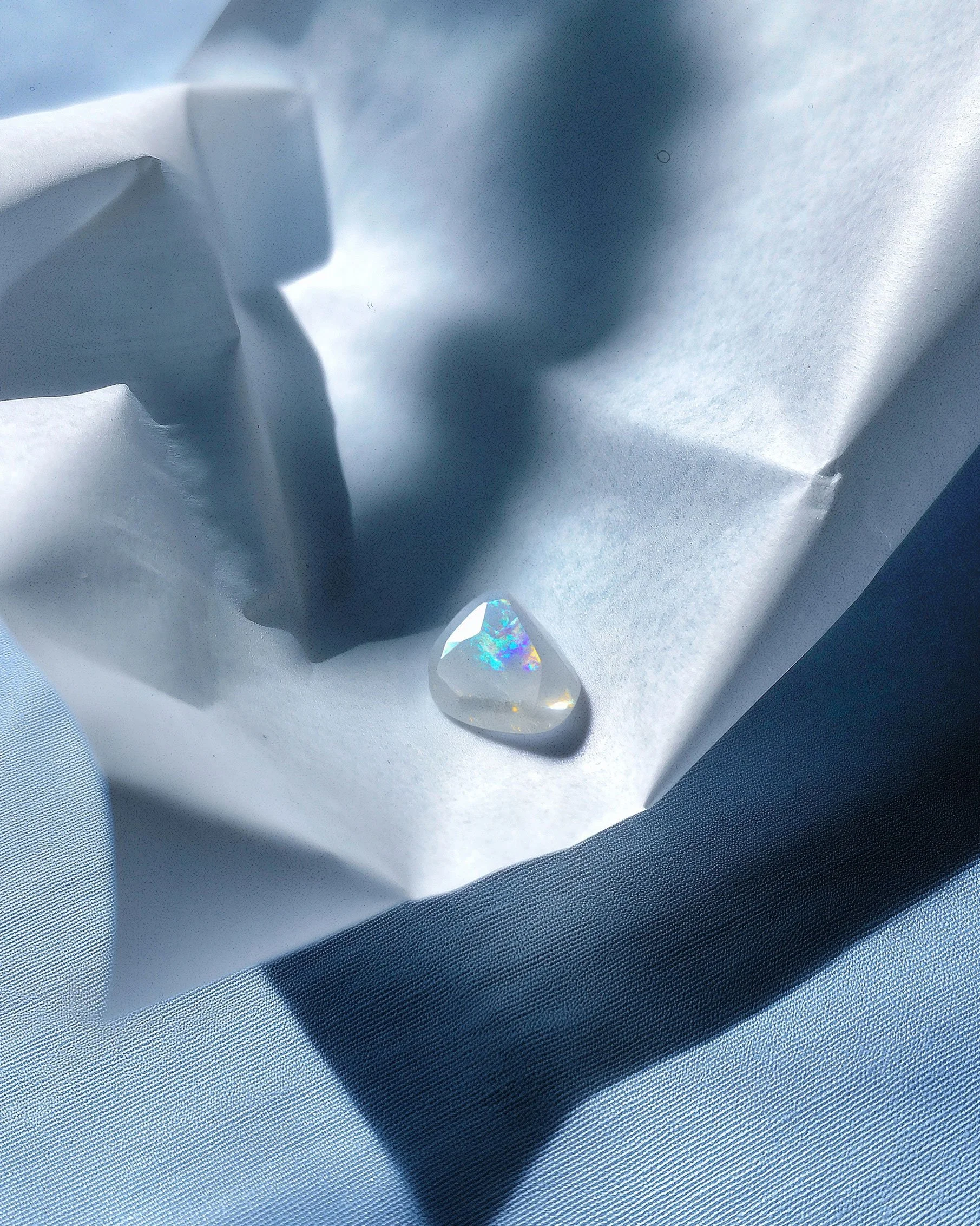 A small, faceted crystal with rainbow colors on a white tissue paper, with shadows cast on light blue fabric.