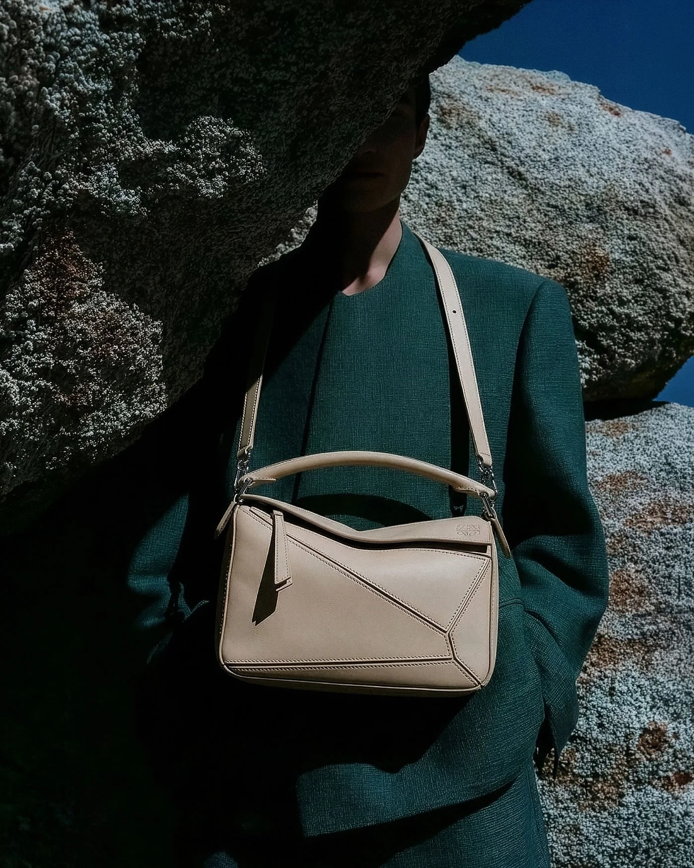 A person wearing a dark green jacket and holding a beige designer handbag, standing next to large textured rocks against a clear blue sky.