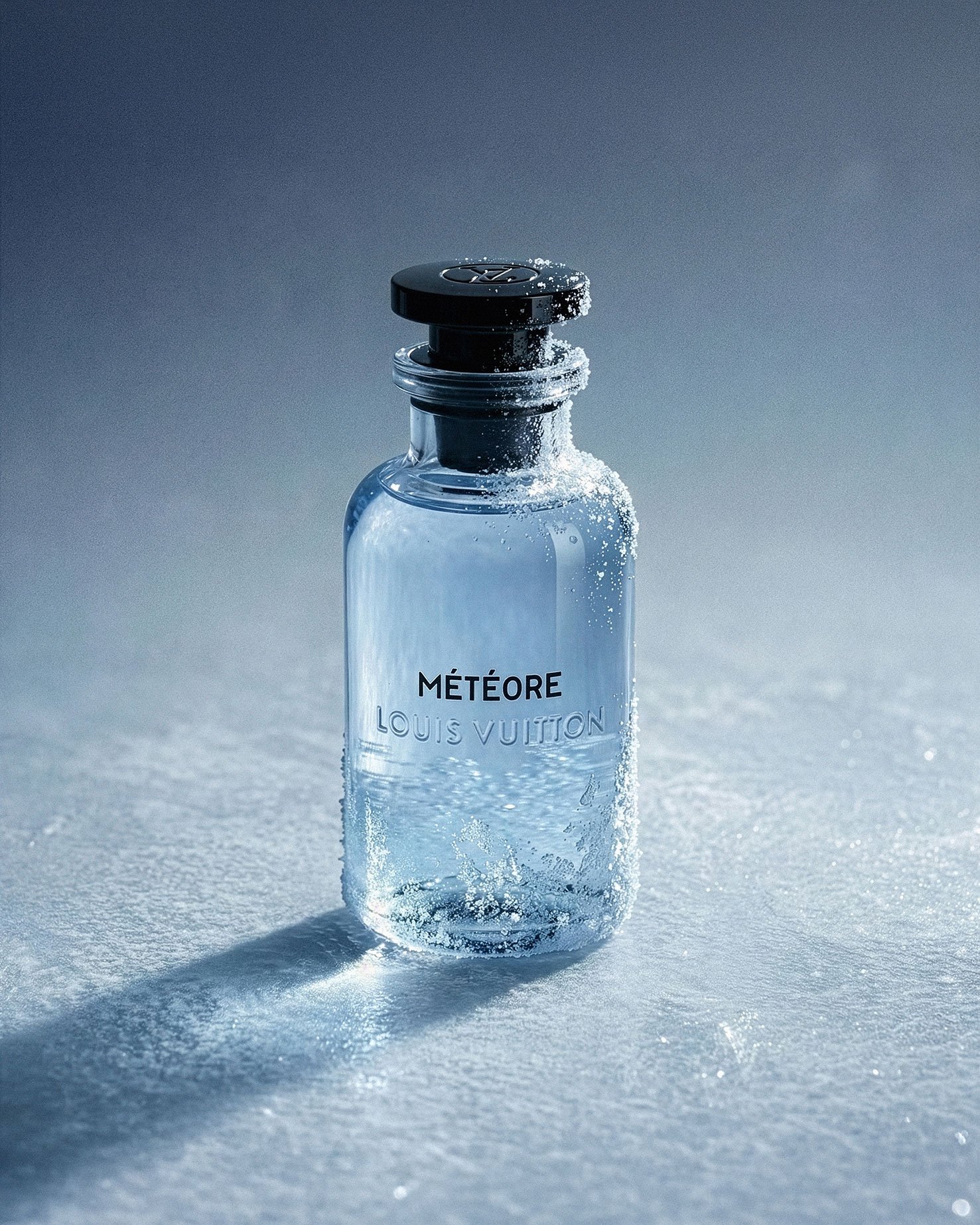 A glass bottle of Louis Vuitton MÉTÉORE perfume with snow on and around it, casting a shadow on a snow-covered surface under a gradient gray background.