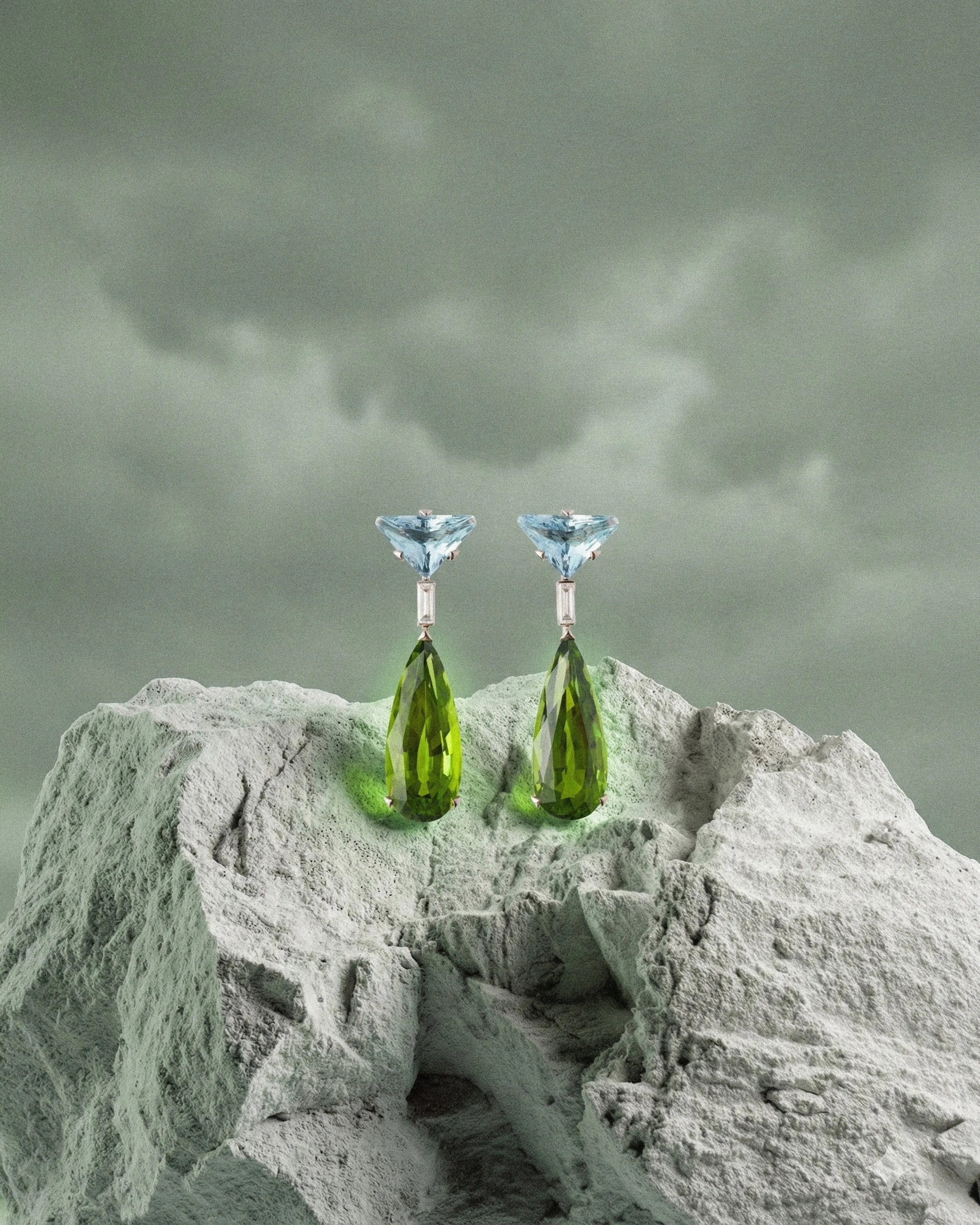 A pair of earrings with large green teardrop-shaped gemstones and smaller blue triangular gemstones, displayed on a rough, white rock with a stormy gray sky in the background.