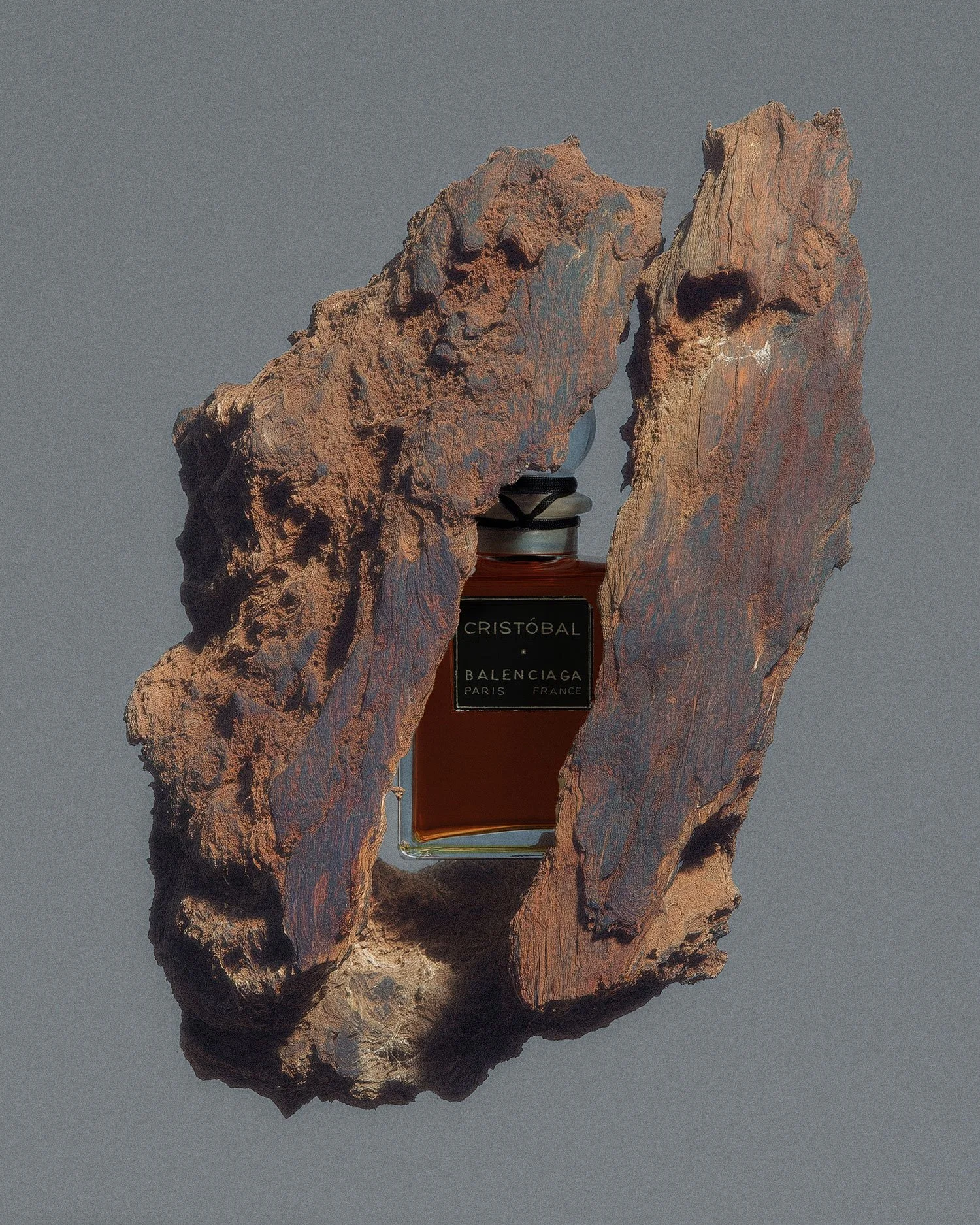 A perfume bottle labeled 'Cristobal Balenciaga Paris France' encased in a rough, natural wood frame against a neutral background.