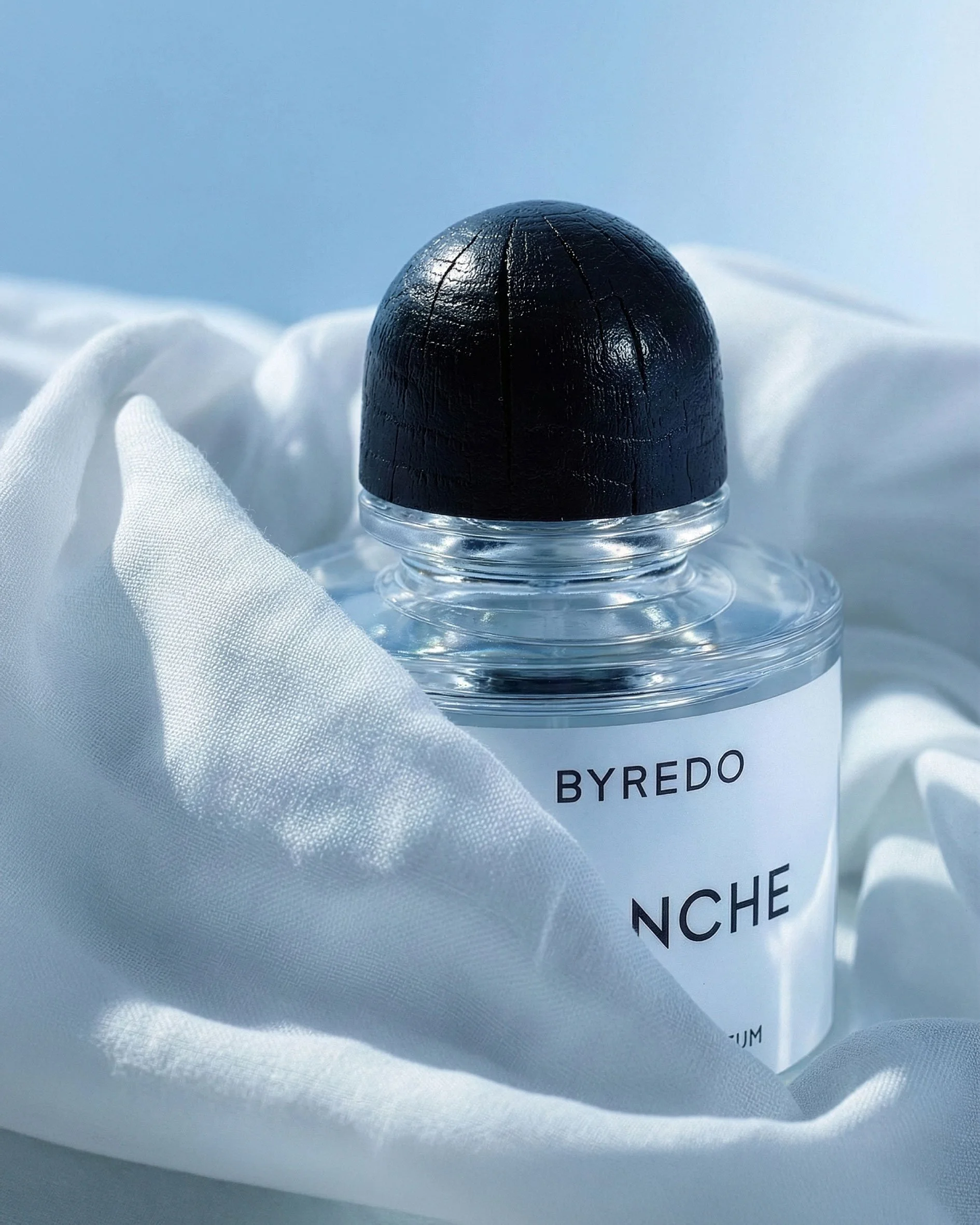 A perfume bottle with a clear glass body, a black rounded cap, and a white label displaying 'BYREDO NCHE' rests on white fabric with a soft, smooth background.