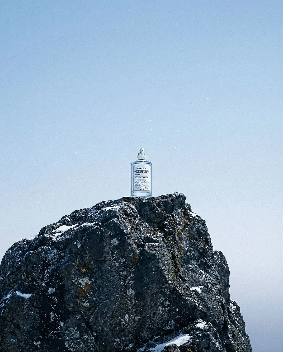 A spray bottle labeled 'REPLICA' placed on top of a large, dark, rocky mountain with patches of snow, against a clear, blue sky.