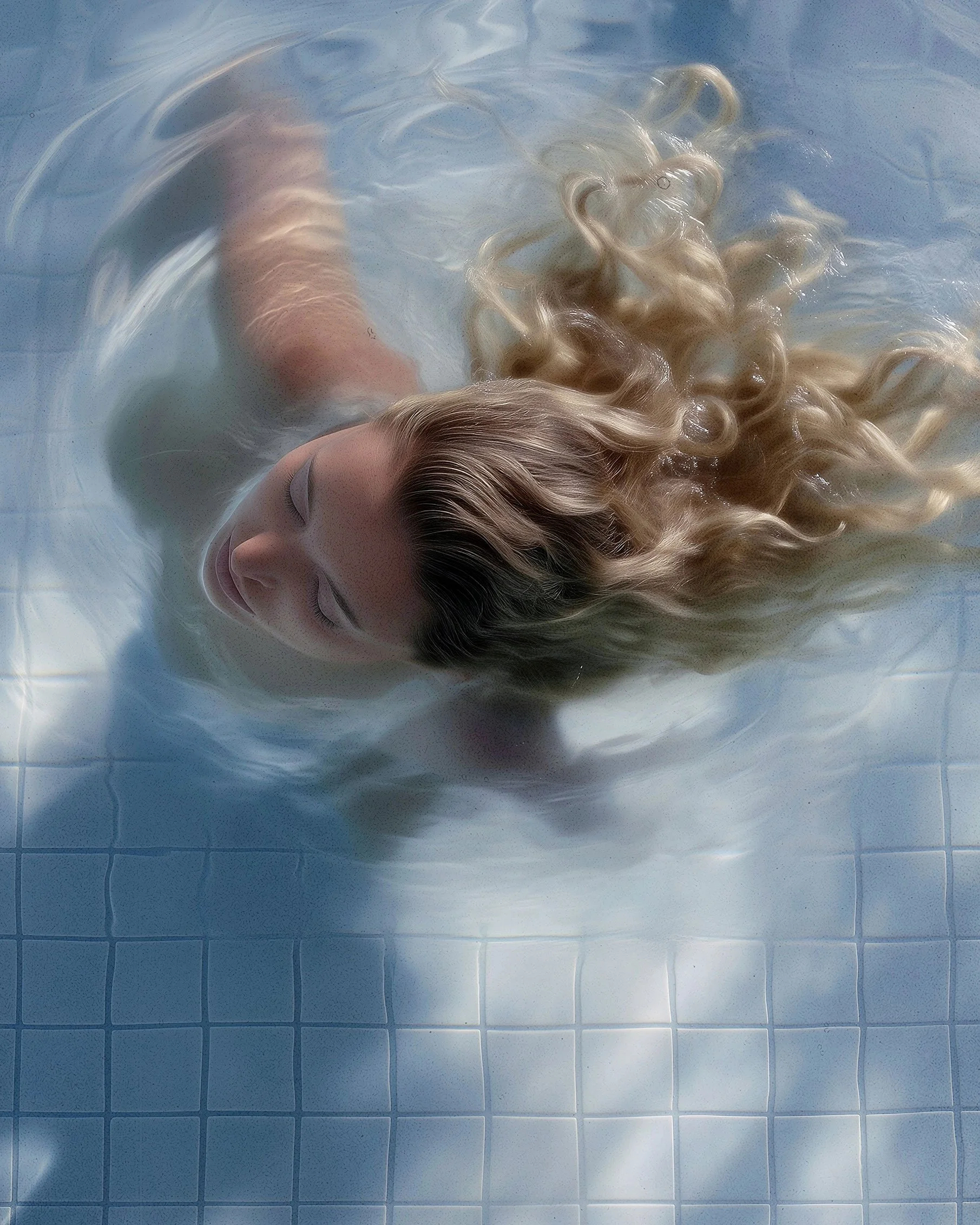 A woman with long blonde curly hair floating on her back in a swimming pool with light blue tiles, eyes closed, and a calm expression.