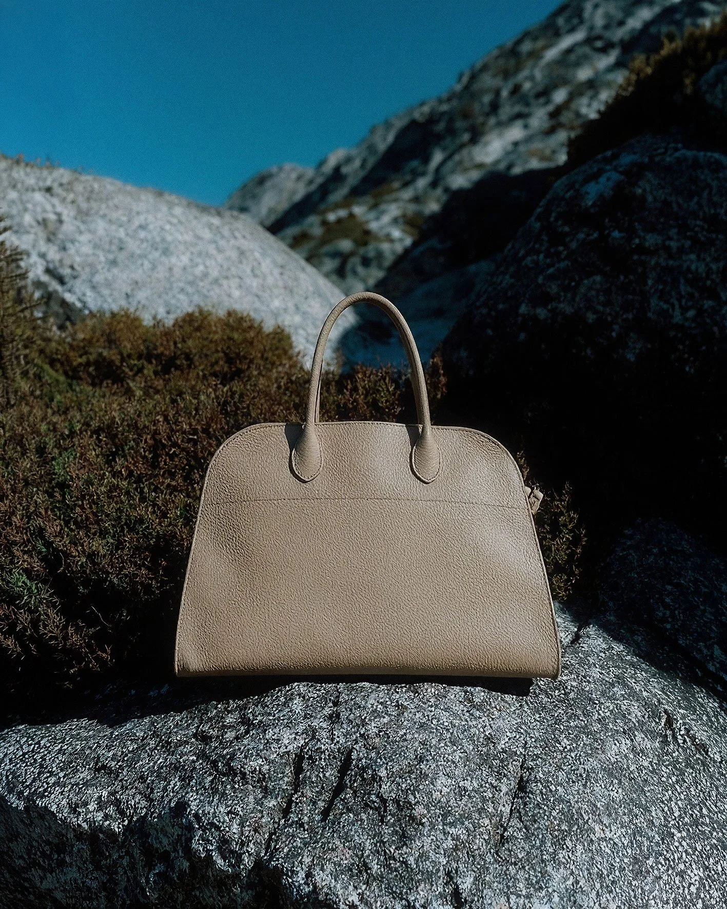 A beige leather handbag resting on a large gray rock outdoors with mountains and blue sky in the background.