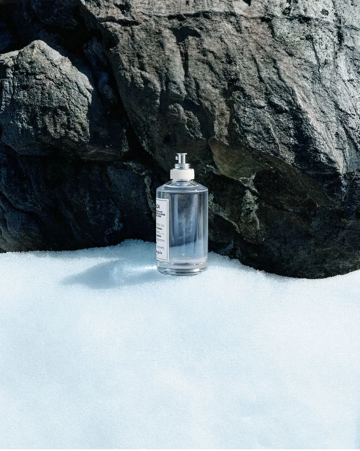 A clear plastic pump bottle filled with hand sanitizer on white sandy ground, with large dark rocks in the background.