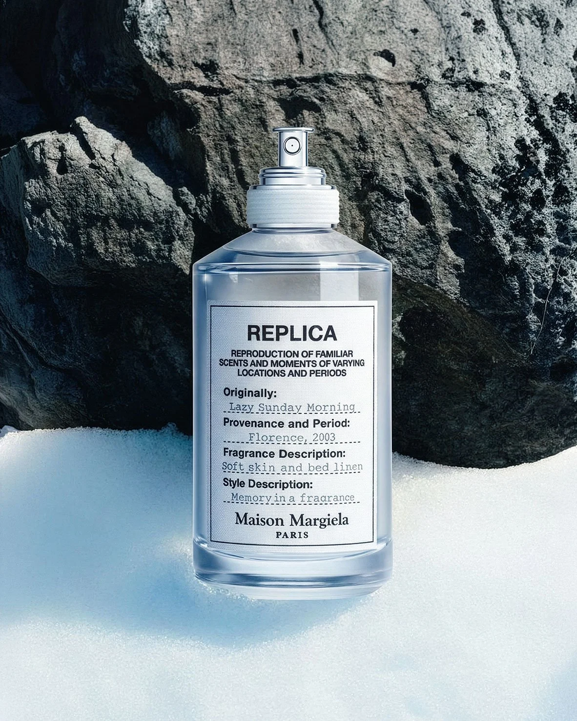 A clear spray bottle labeled 'REPLICA,' placed on snow with rocks in the background.