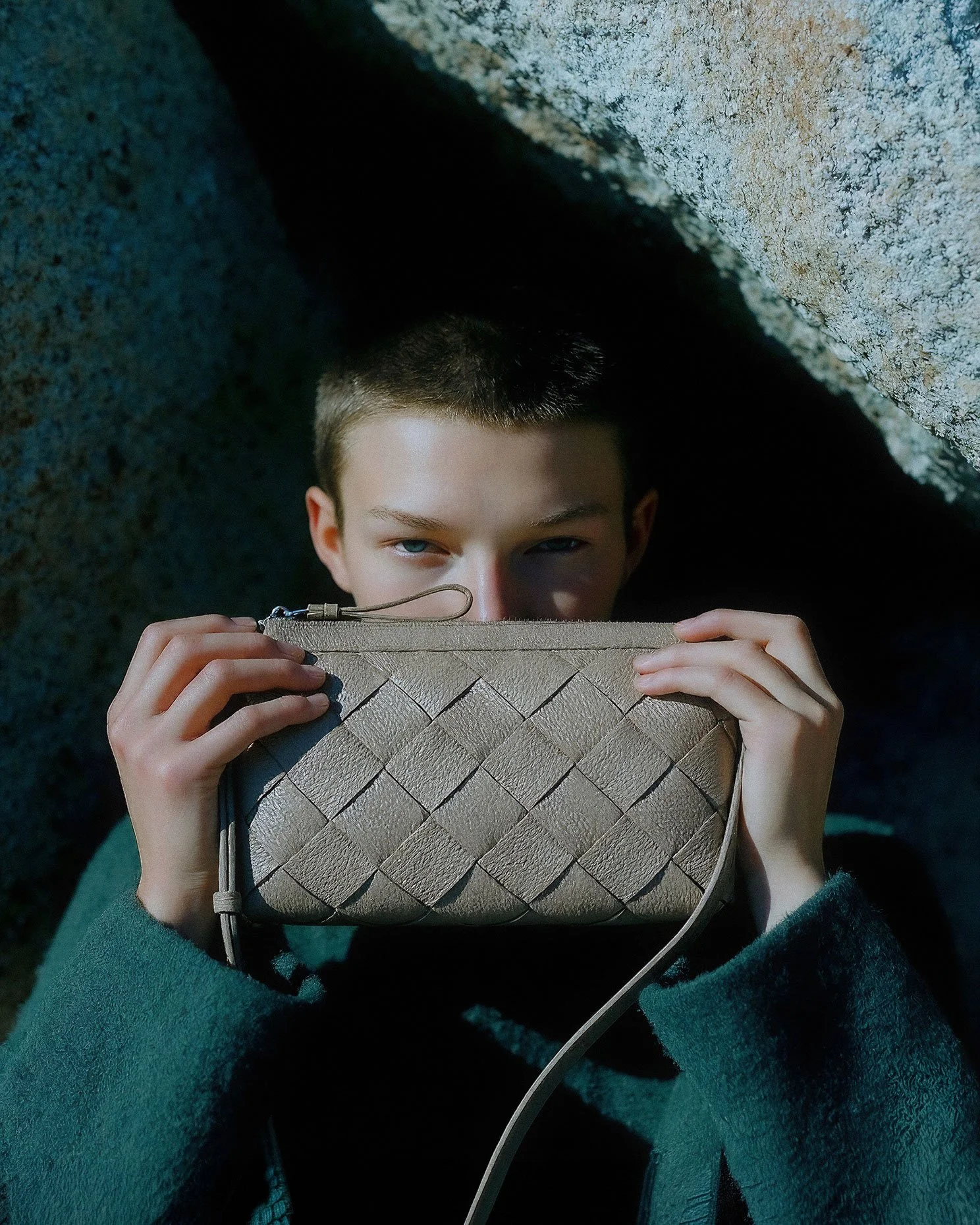 A young person with short hair holding a woven leather clutch in front of their face, peeking over the top.