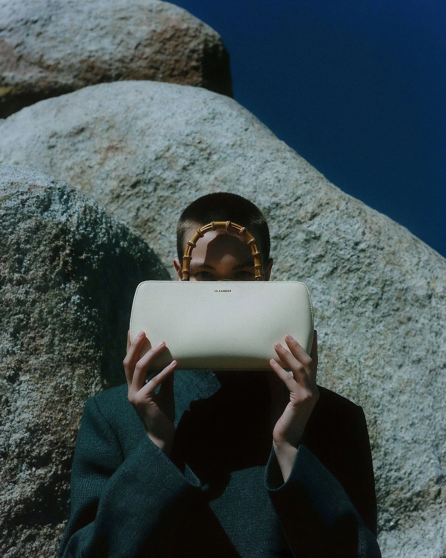 Person with short hair holding a white Jil Sander handbag, standing outdoors in front of large rocks, with their face partially obscured.