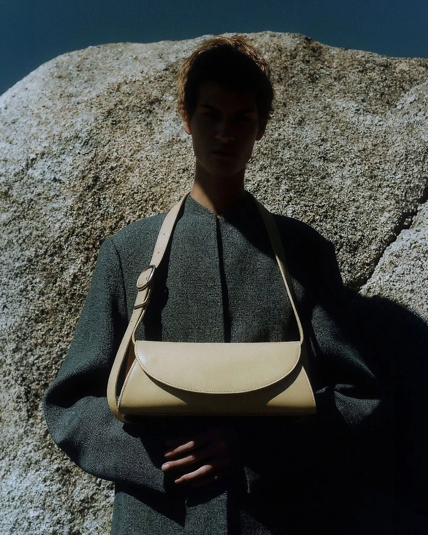 Person standing in front of a large rock with shadowed face, wearing a dark long-sleeve top and carrying a beige shoulder bag, with clear blue sky in the background.