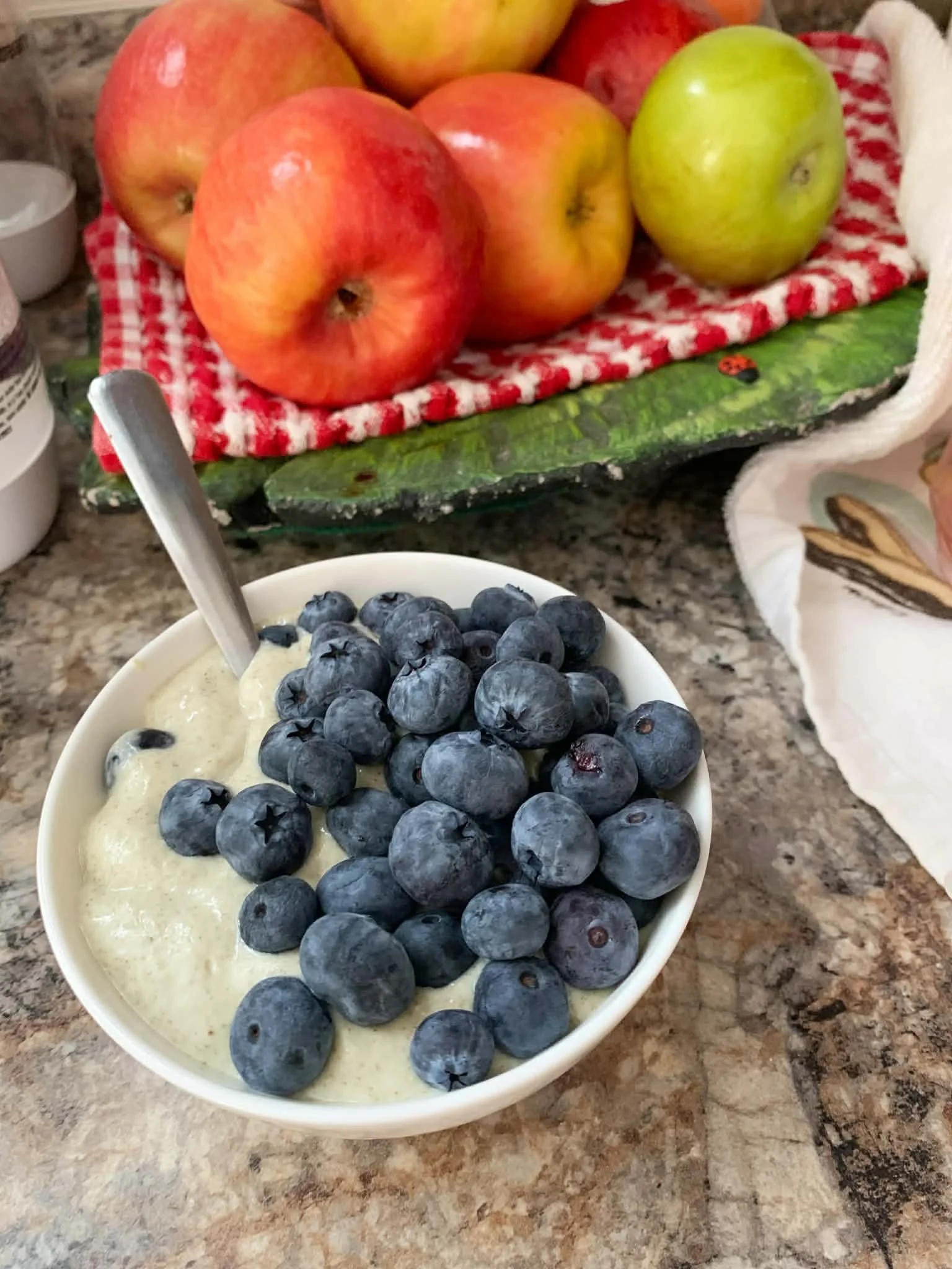 small white bowl with light green chia pudding, topped with lots of blueberries, next to a bowl of apples