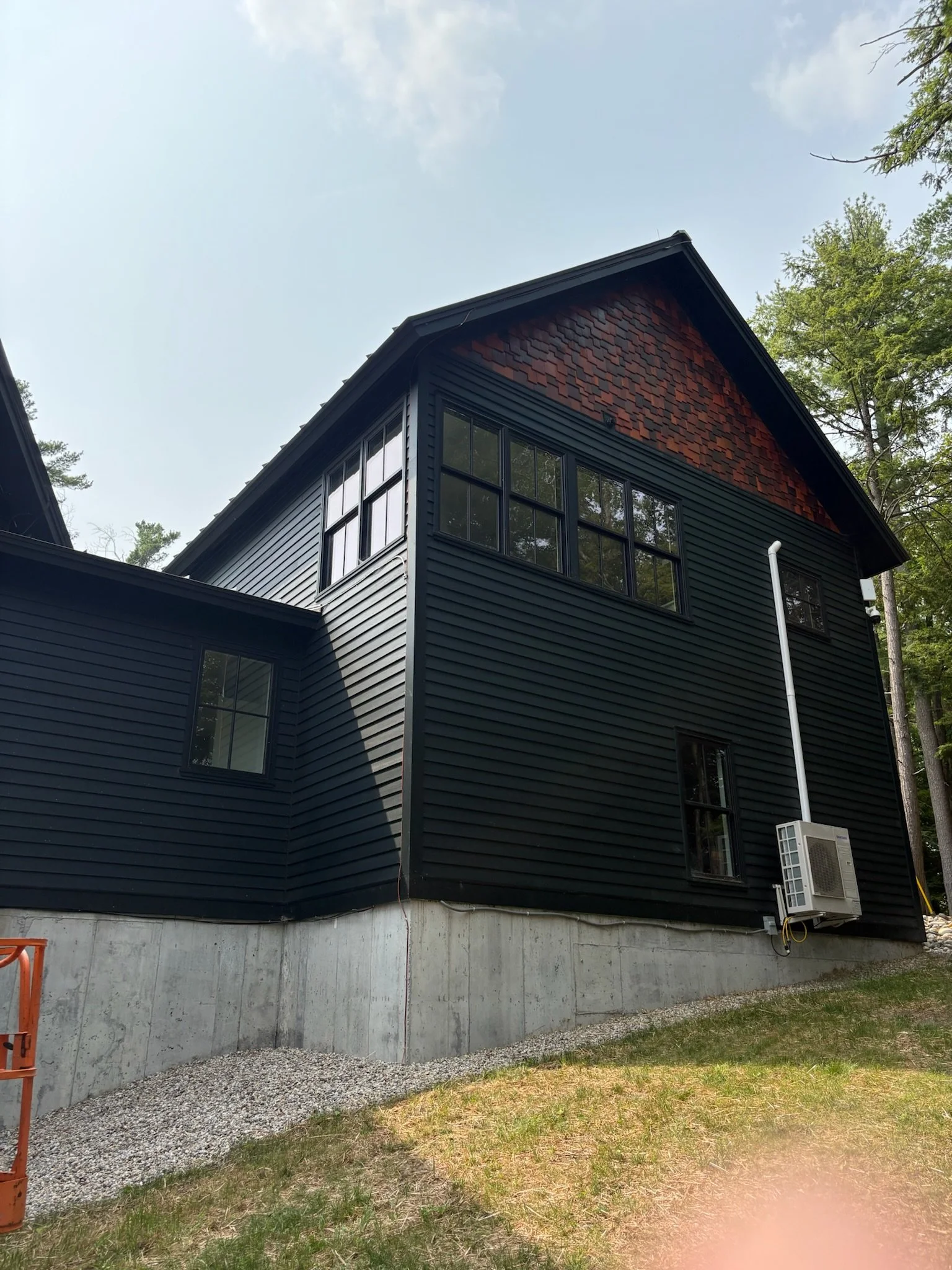 Back of a black house with large windows, a red shingle gable, and an air conditioning unit on the side, set in a wooded area with trees and a partly cloudy sky.