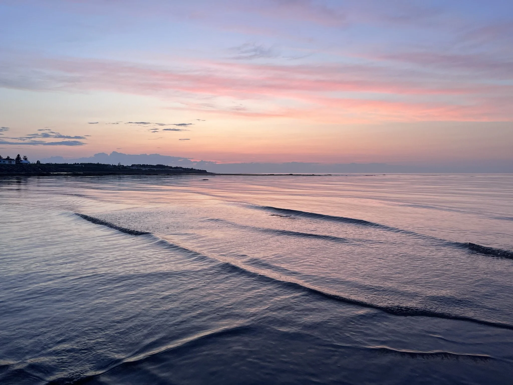 Calm beach at sunset with pink and purple sky, gentle waves, and dark silhouette of land in the background.