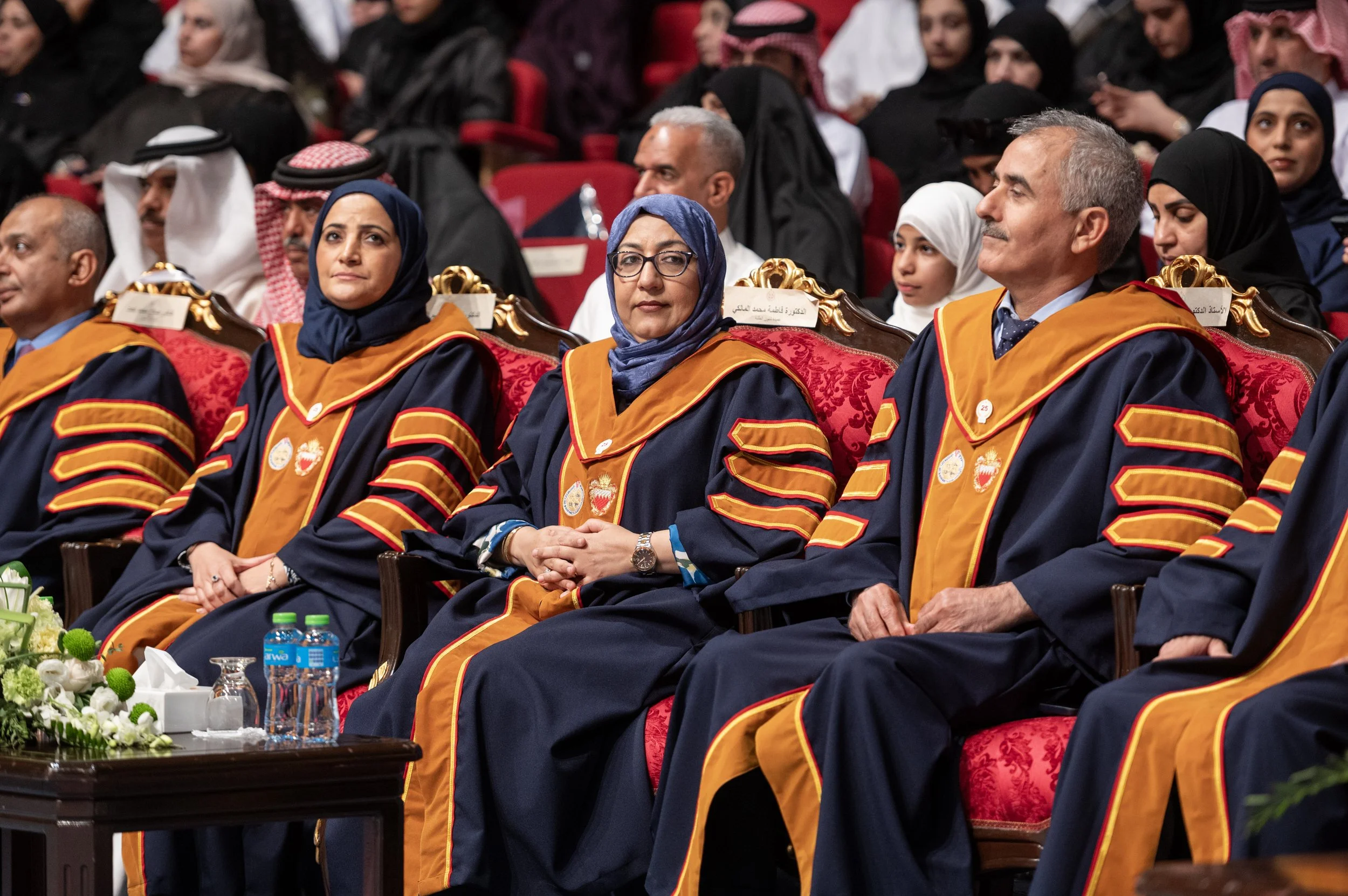 Group of people sitting in an auditorium at a graduation ceremony, wearing academic regalia with gold and black robes.