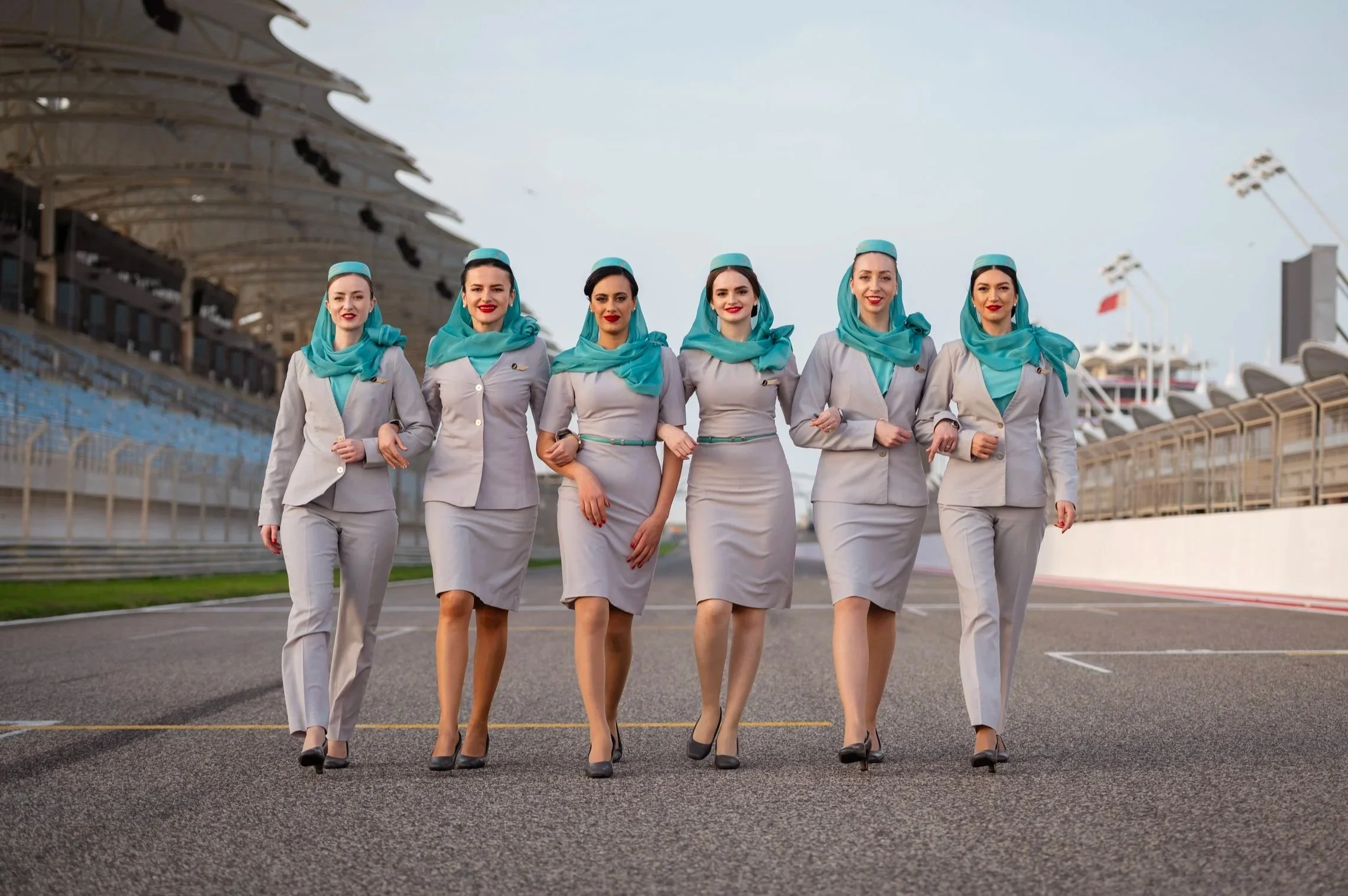 Six women in flight attendant uniforms, wearing matching grey suits with teal scarves and hats, walking on a race track at an airport, smiling at the camera.