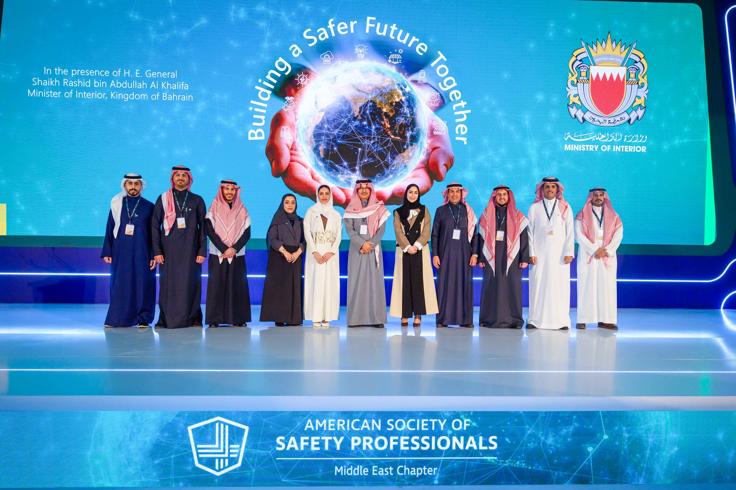 Group of men and women standing on stage for a photo at a conference. The backdrop has a globe with hands around it and the phrase "Building a Safer Future Together." The event is organized by the Ministry of Interior in Bahrain, and participants are