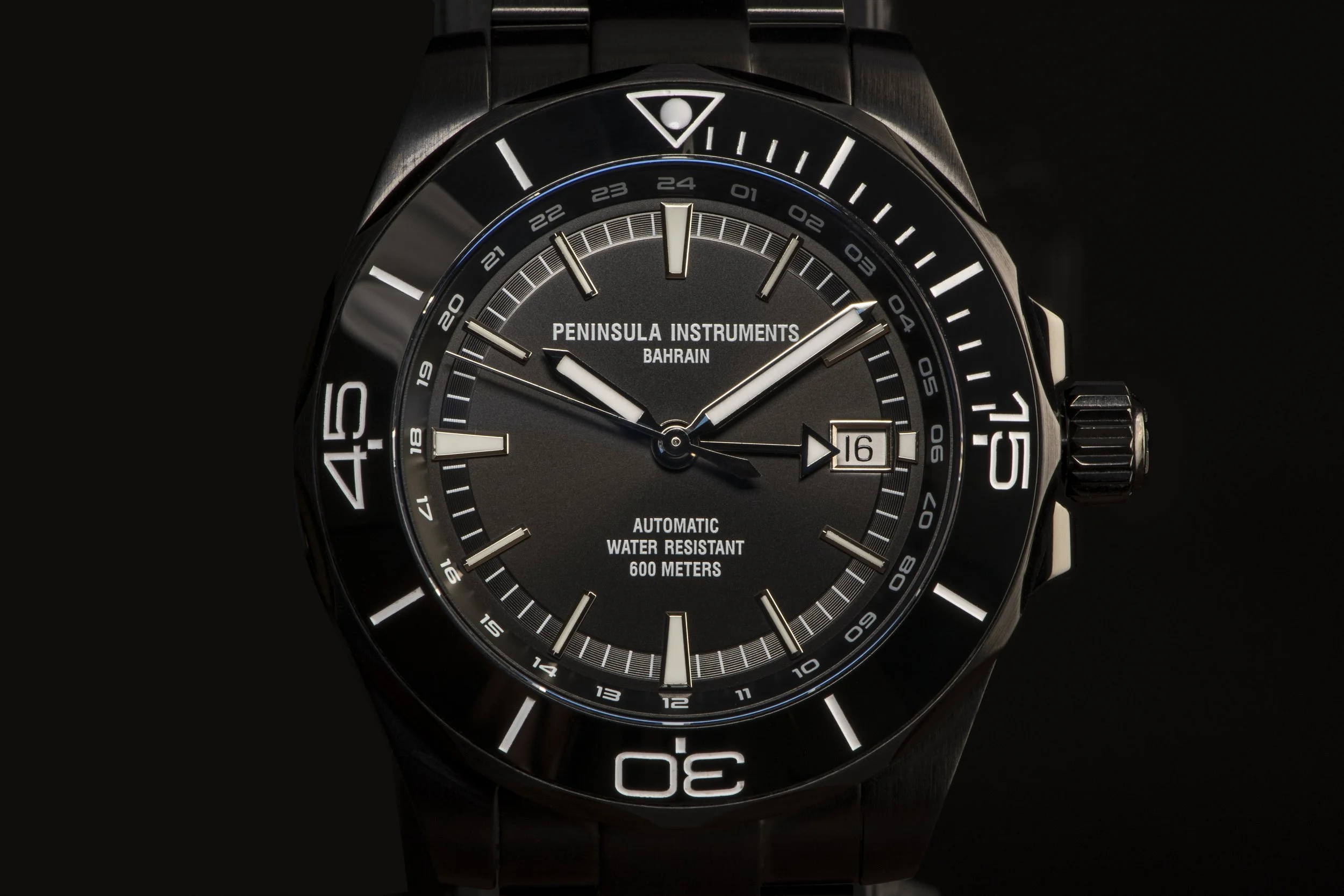 Close-up of a black and silver wristwatch with a black dial, white markers, and a date window at 3 o'clock. The watch has a rotating bezel with minute markings and is labeled Peninsula Instruments Bahrain, water resistant to 600 meters.