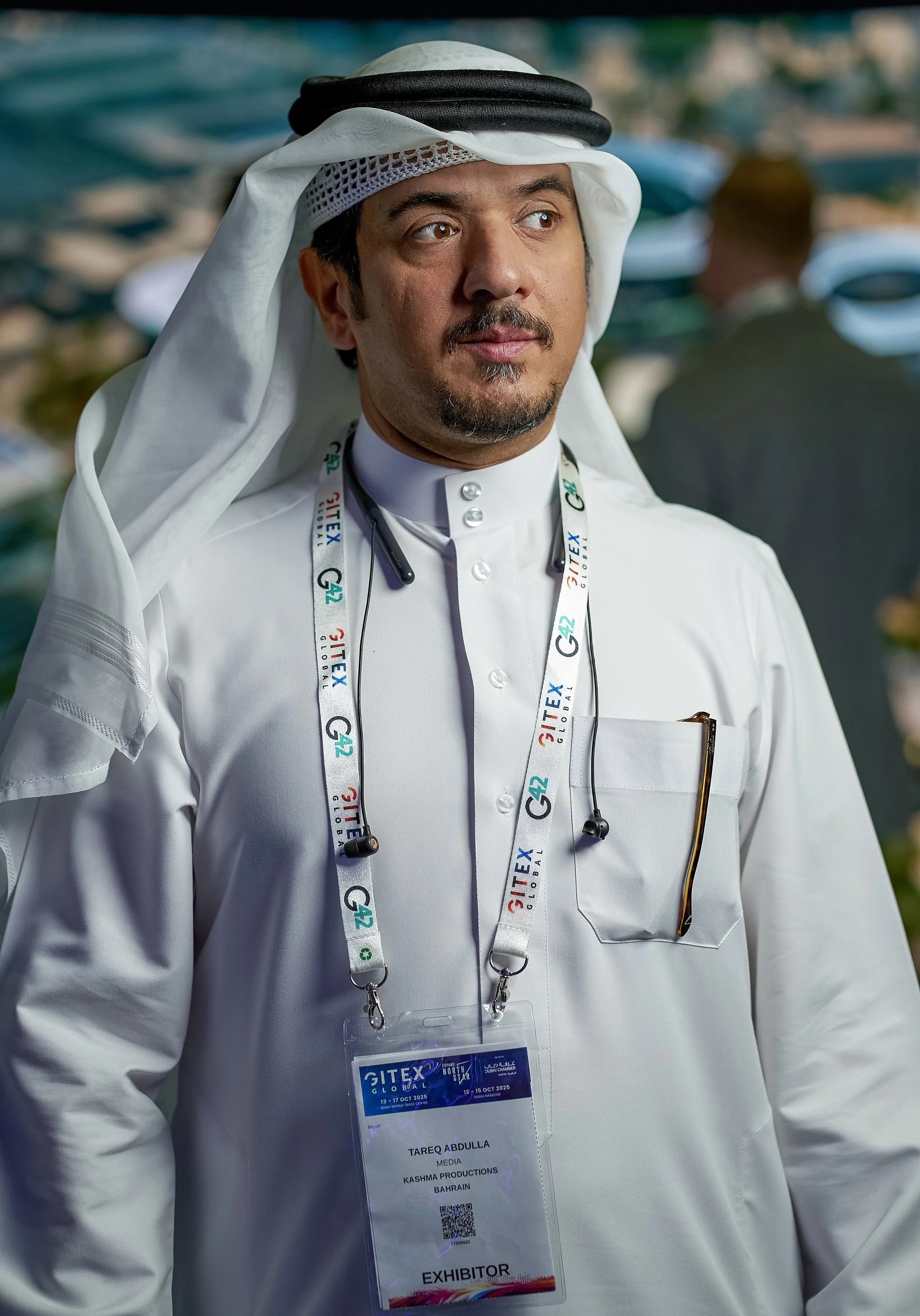 Man wearing traditional Middle Eastern attire, including a white thobe and a head covering, with an exhibitor badge at a conference, with blurred background of water and boats.
