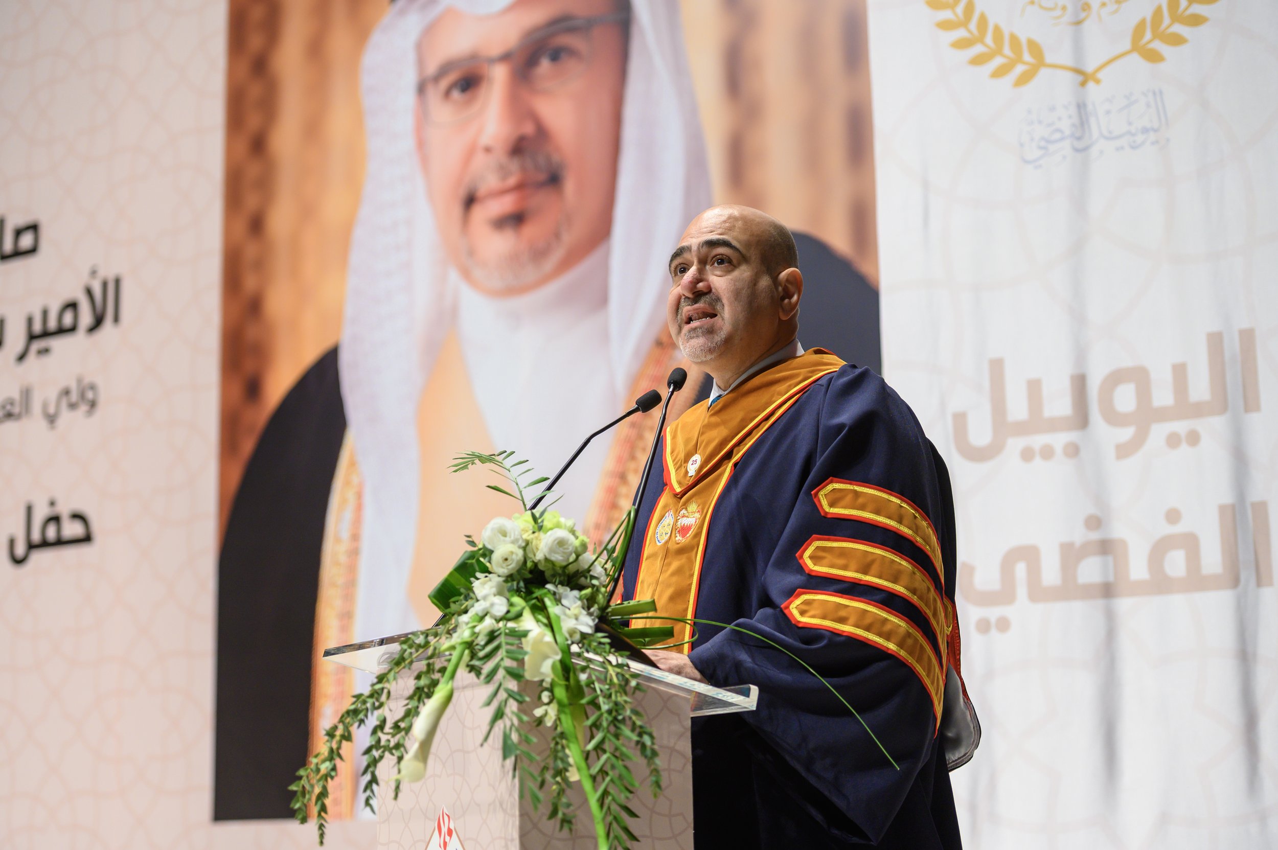 A man dressed in academic regalia giving a speech at a podium decorated with a bouquet of white flowers, with a large poster of himself and Arabic text in the background.