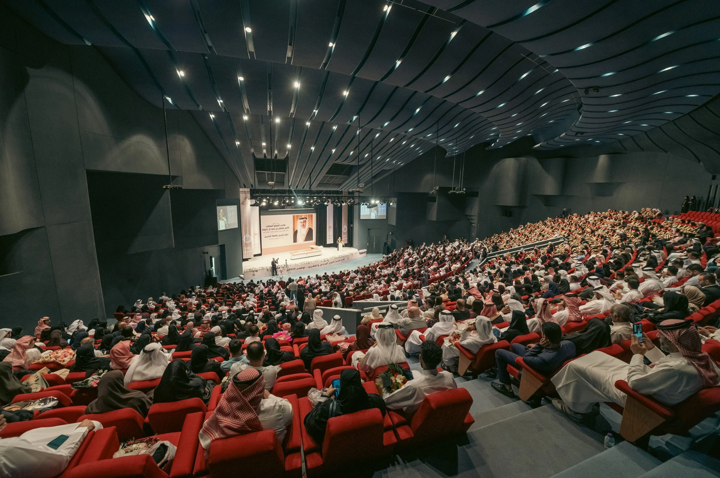A large auditorium filled with audience members wearing traditional Middle Eastern clothing, attending a conference or presentation. The stage has screens displaying a speaker, and there are decorations with Arabic writing and images of a man on the 