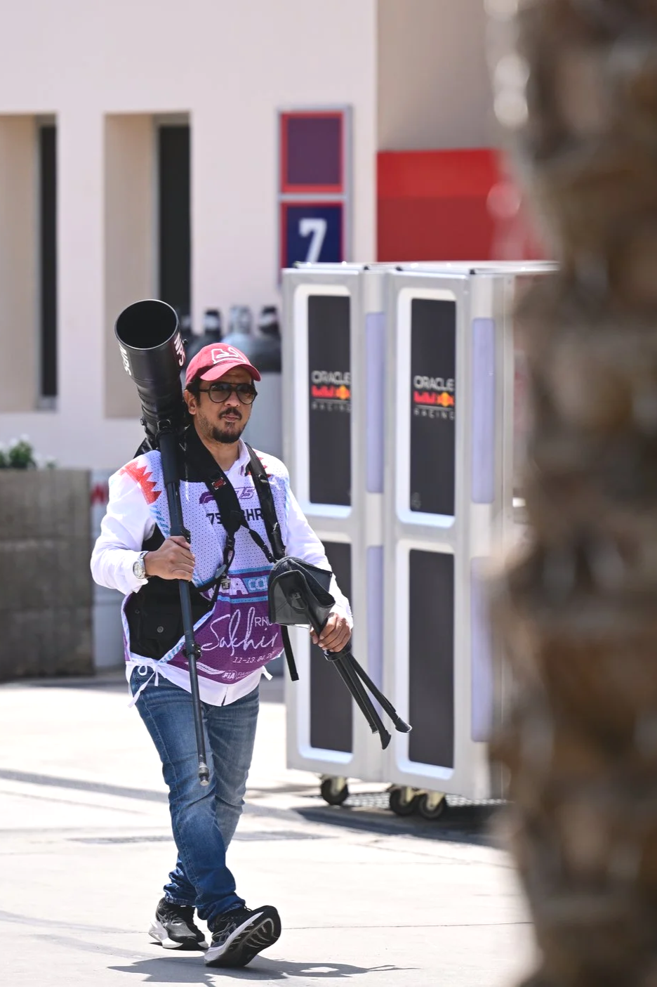 Man wearing a red cap, sunglasses, and a white and purple sports jersey, carrying a large camera lens and tripod, walking outdoors near a building with a sign reading '7' and orange Oracle advertising posters.