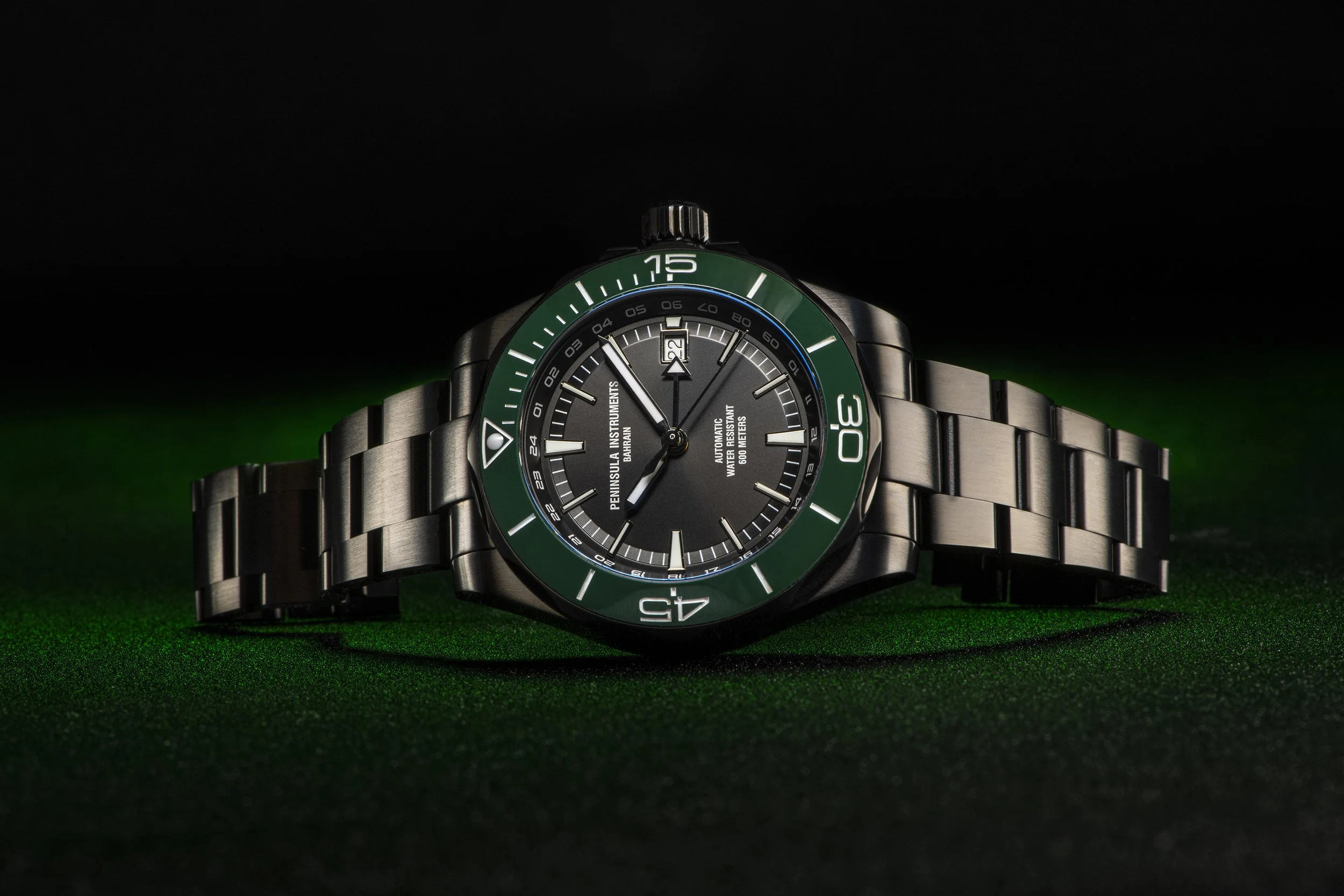 Silver wristwatch with a black and green bezel and a metal bracelet, laid on a green surface with a dark background.