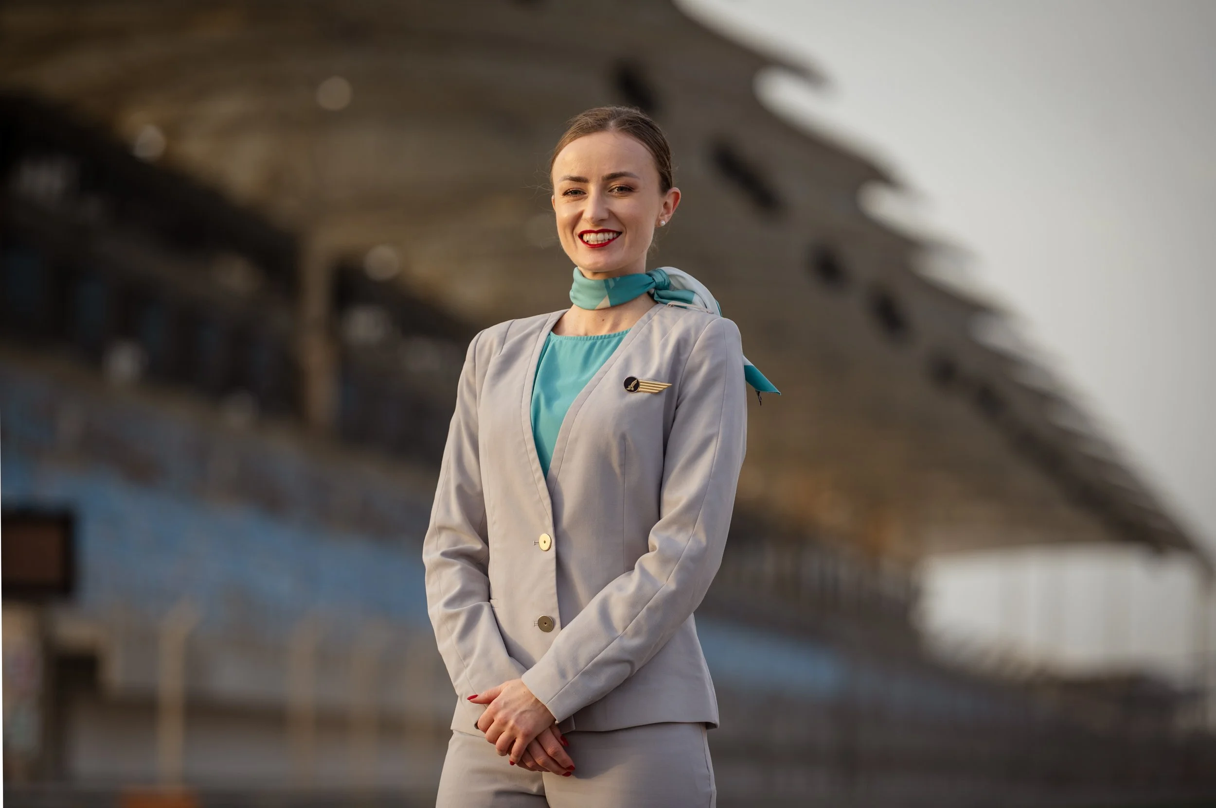 A smiling woman dressed in a vintage flight attendant uniform with a teal scarf tied around her neck and a winged badge on her blazer, standing outdoors in front of a blurred modern building, likely at an airport.