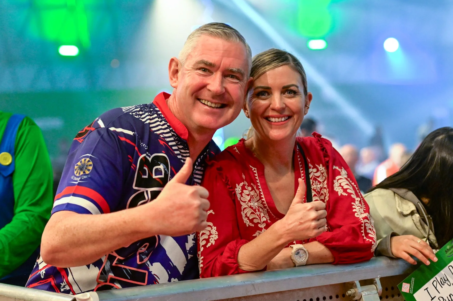 A smiling man and woman at a crowded event, both giving a thumbs-up gesture. The man is wearing a colorful sports shirt and the woman is wearing a red embroidered blouse. Bright lights and other people are visible in the background.