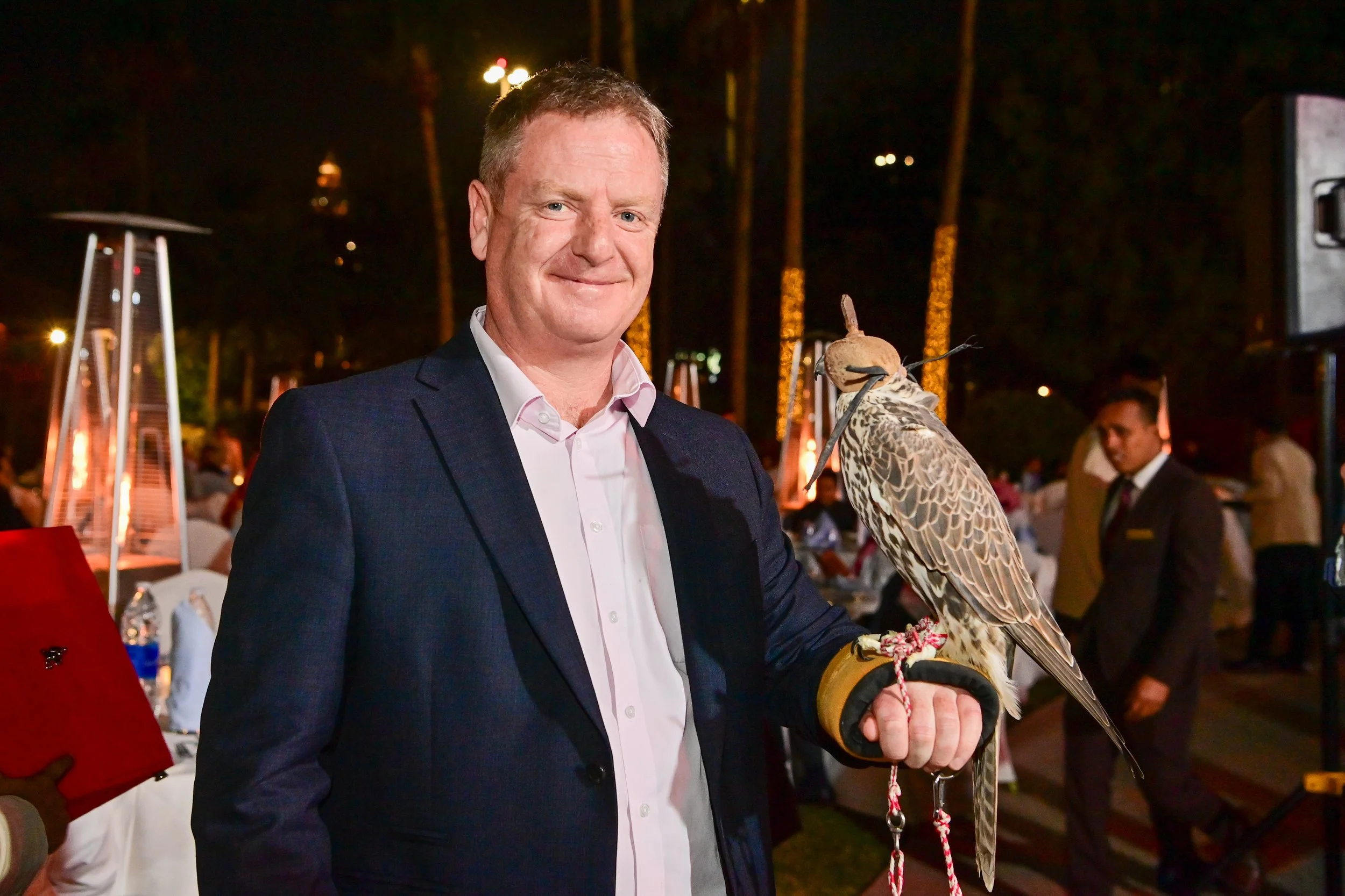 A man in a suit holding a falcon on his gloved hand at an outdoor event at night.