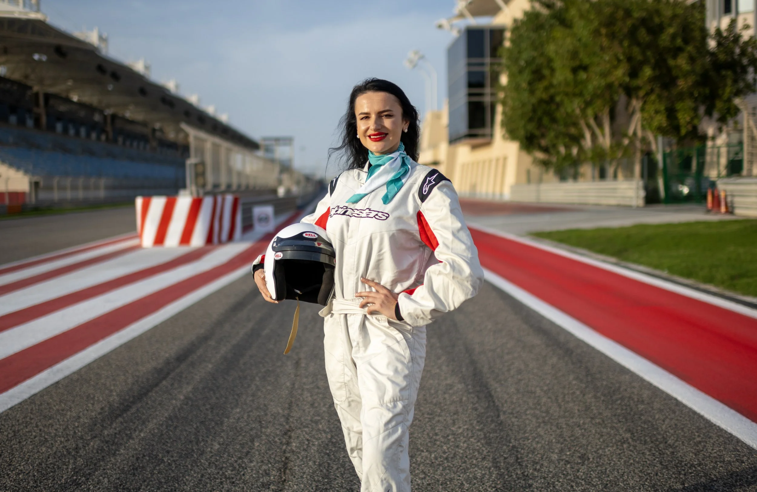A woman in a white racing suit standing on a race track, holding a helmet, with her hands on her hips, smiling at the camera.