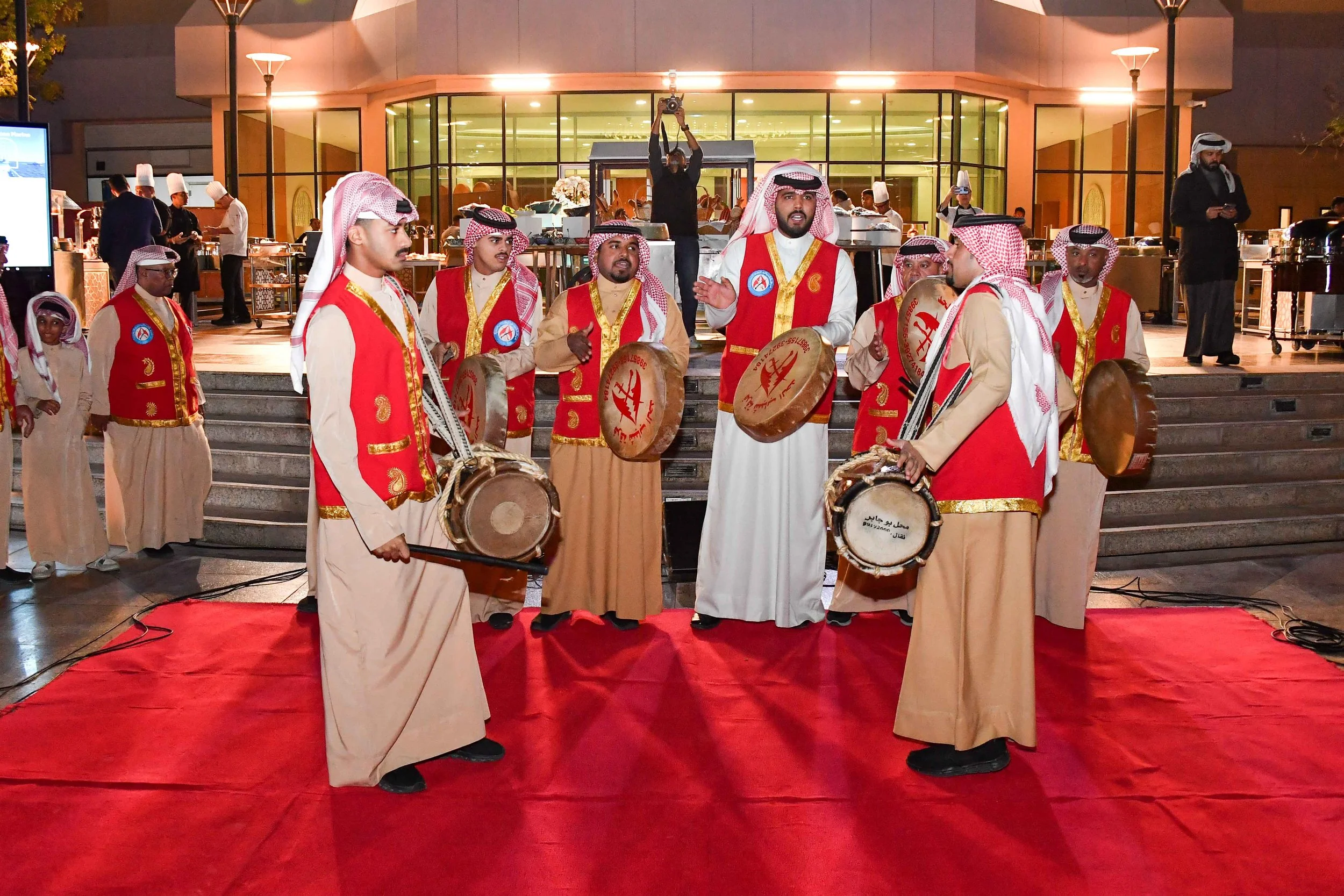 A group of men in traditional Middle Eastern attire performing a drum circle on a red carpet outside a modern building at night, with chefs and other people in the background.