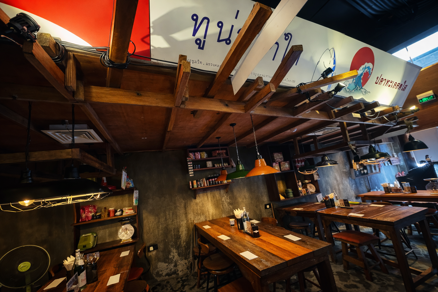 Interior of a restaurant with wooden tables, colorful pendant lights, shelves with condiments, and signs in a non-English language.