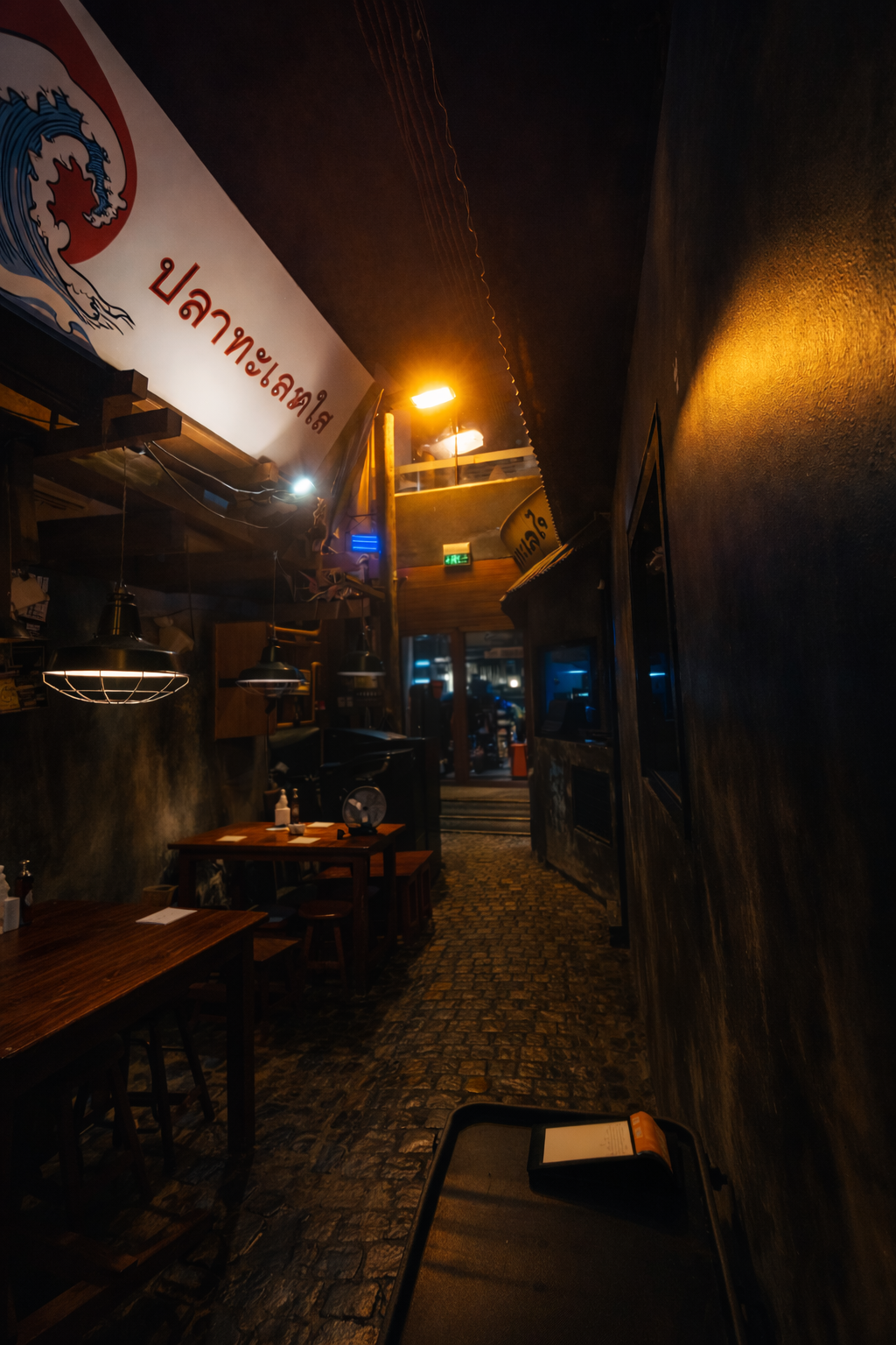 Dimly lit alleyway inside a restaurant or cafe with dark walls, wooden tables, hanging lamps, and a cobblestone floor, illuminated by warm yellow and blue lighting.