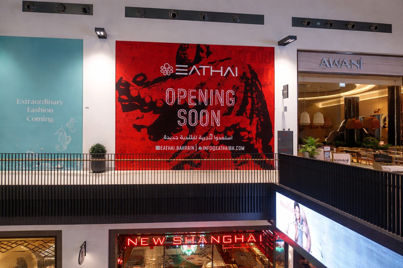 Sign for an upcoming restaurant opening called EATHAI, with the message 'Opening Soon' on a red background inside a shopping mall. The sign includes contact information and social media handles. To the right, part of a restaurant or retail store entrance is visible, with the name 'AWAN' and interior lighting, seating, and decorations.