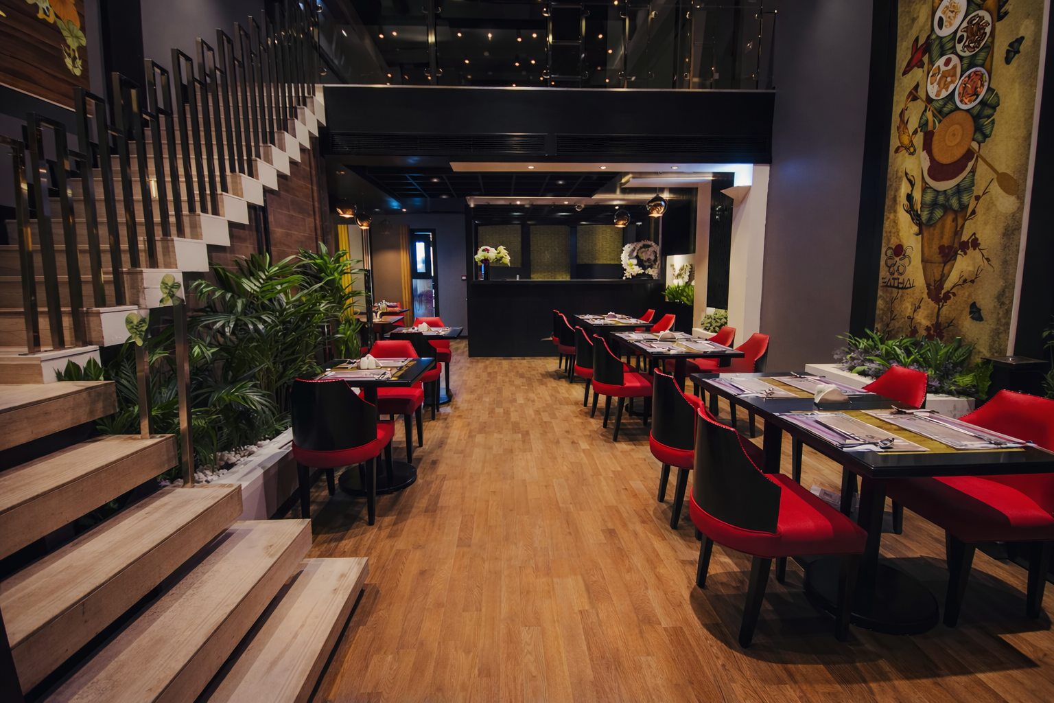 Empty restaurant with red and black chairs, set tables, wooden floors, stairs on the left, plants, and wall art.